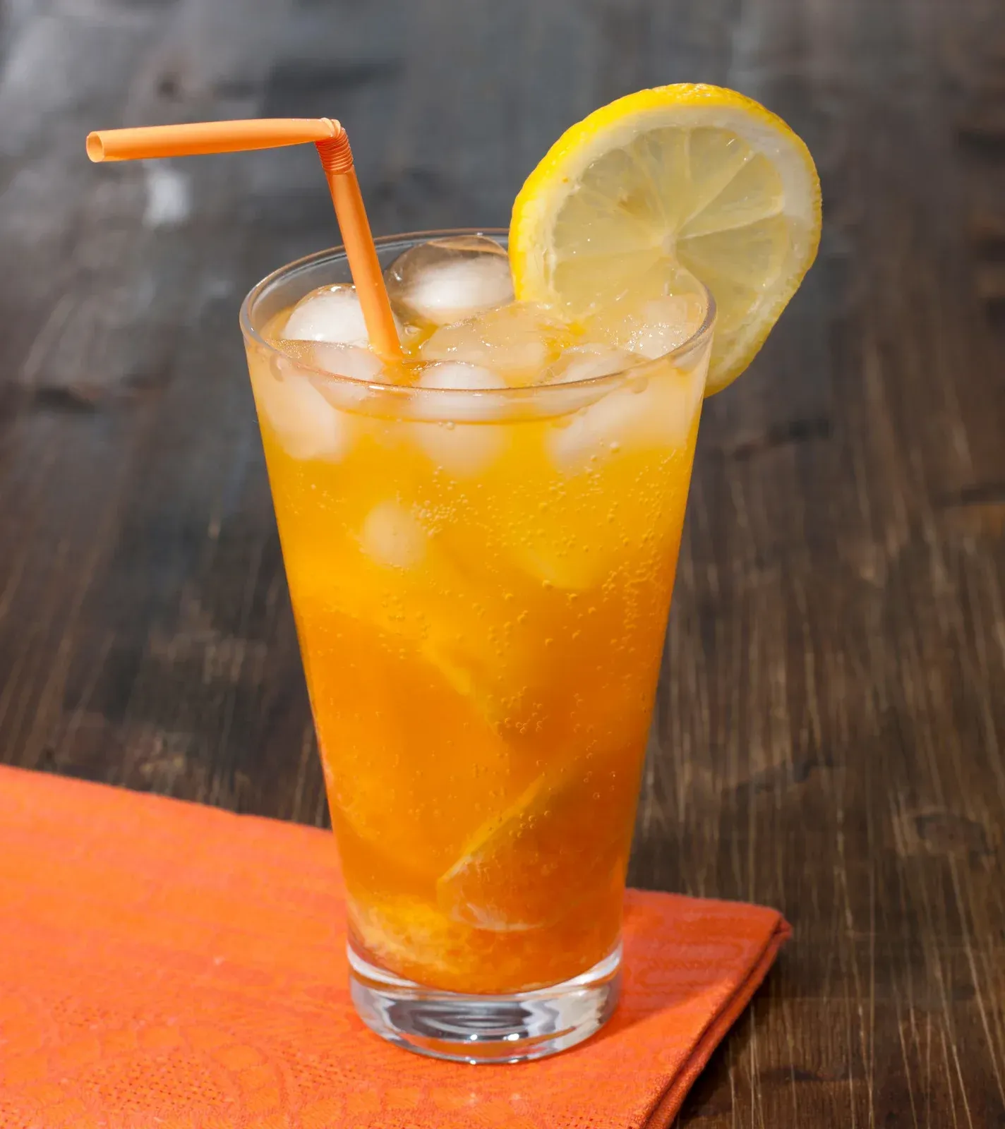 Orange iced drink in a tall glass with a lemon slice and straw on a table