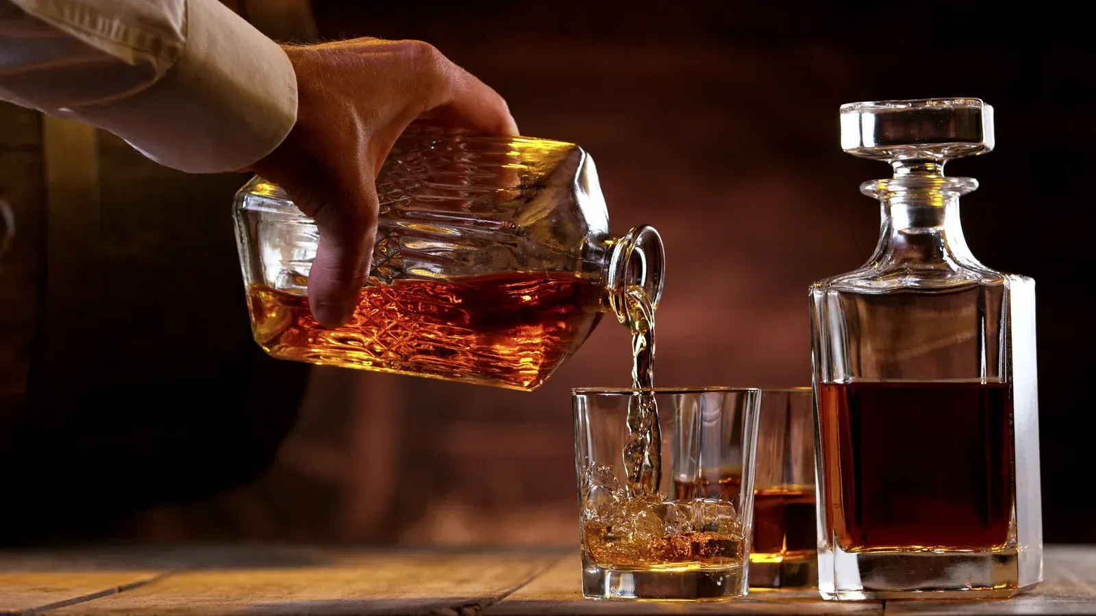 Hand pouring amber liquor from a cut-glass decanter into a tumbler beside a whiskey bottle