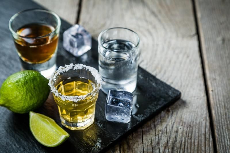 Limes and tequila shots on a black tray with ice, on a rustic wooden table