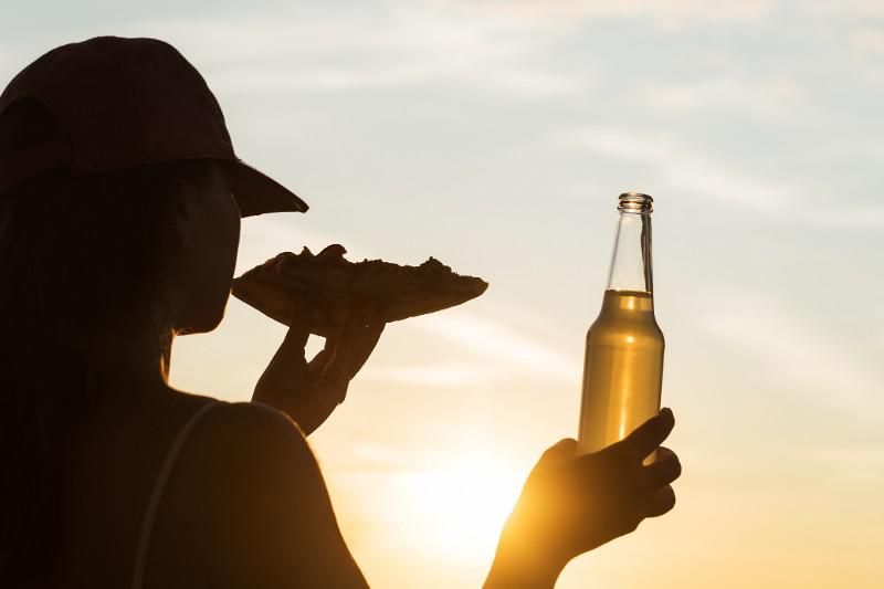 Silhouette holding a slice of pizza and a beer bottle at sunset