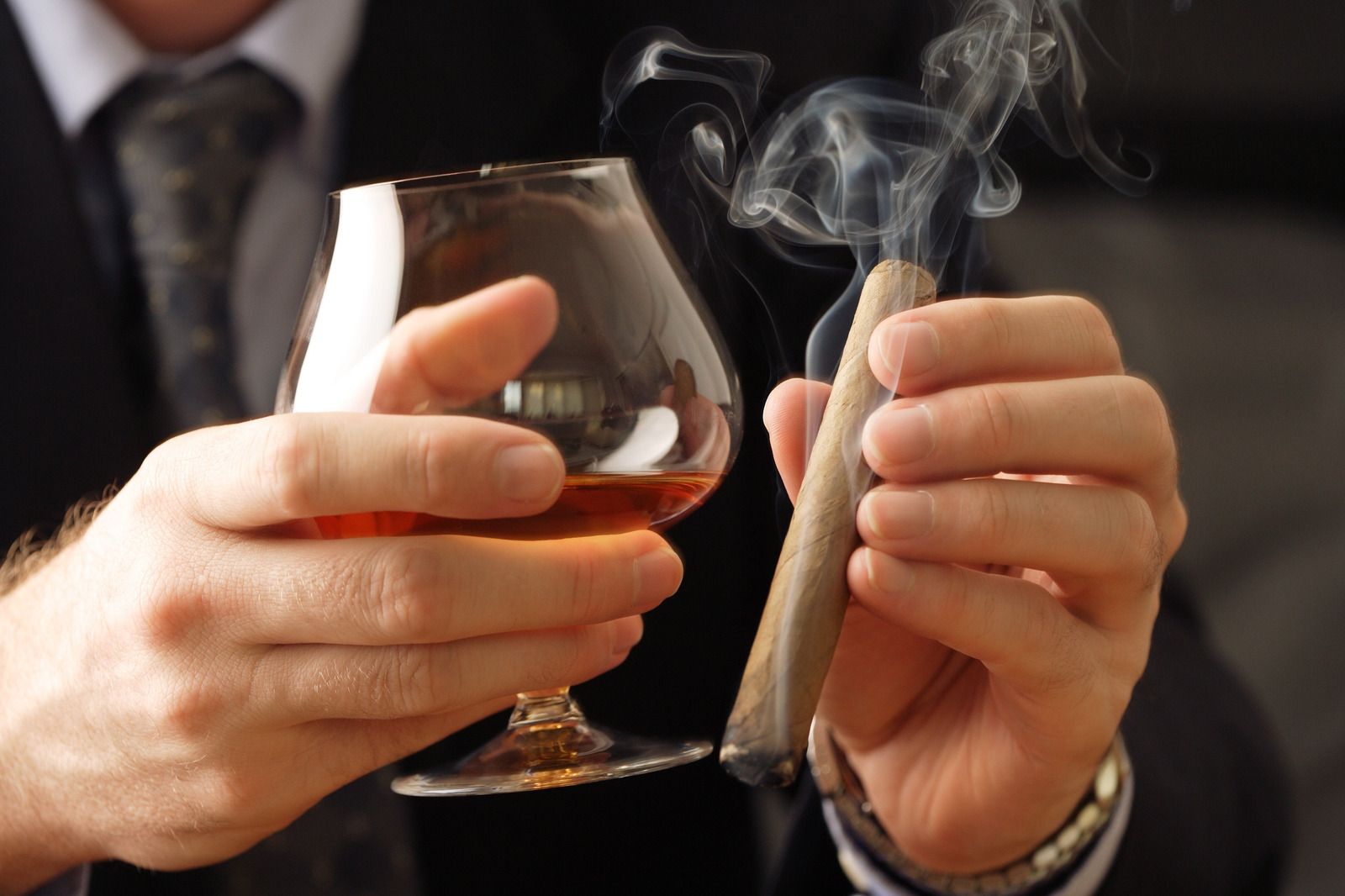 Hands holding a glass of amber liquor and a lit cigar with smoke rising