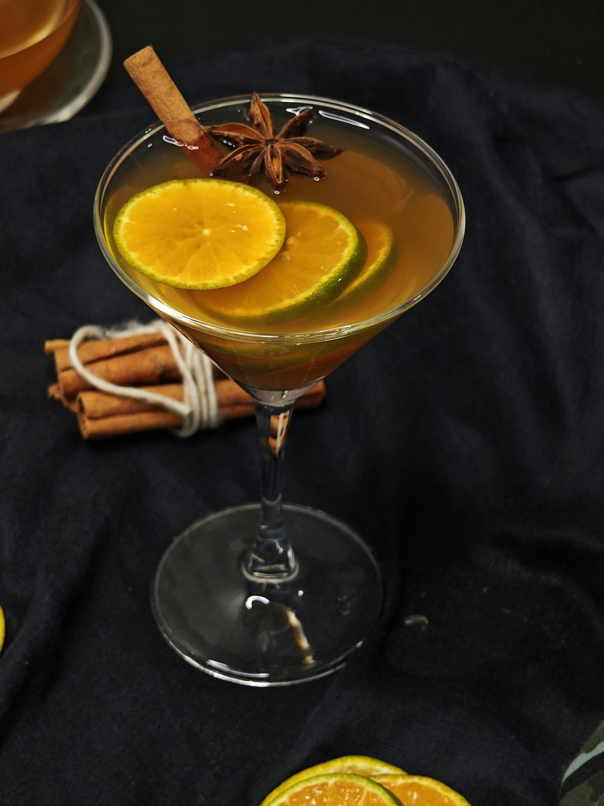 Martini glass with a spiced orange cocktail, orange slices, cinnamon sticks, and star anise on a dark cloth