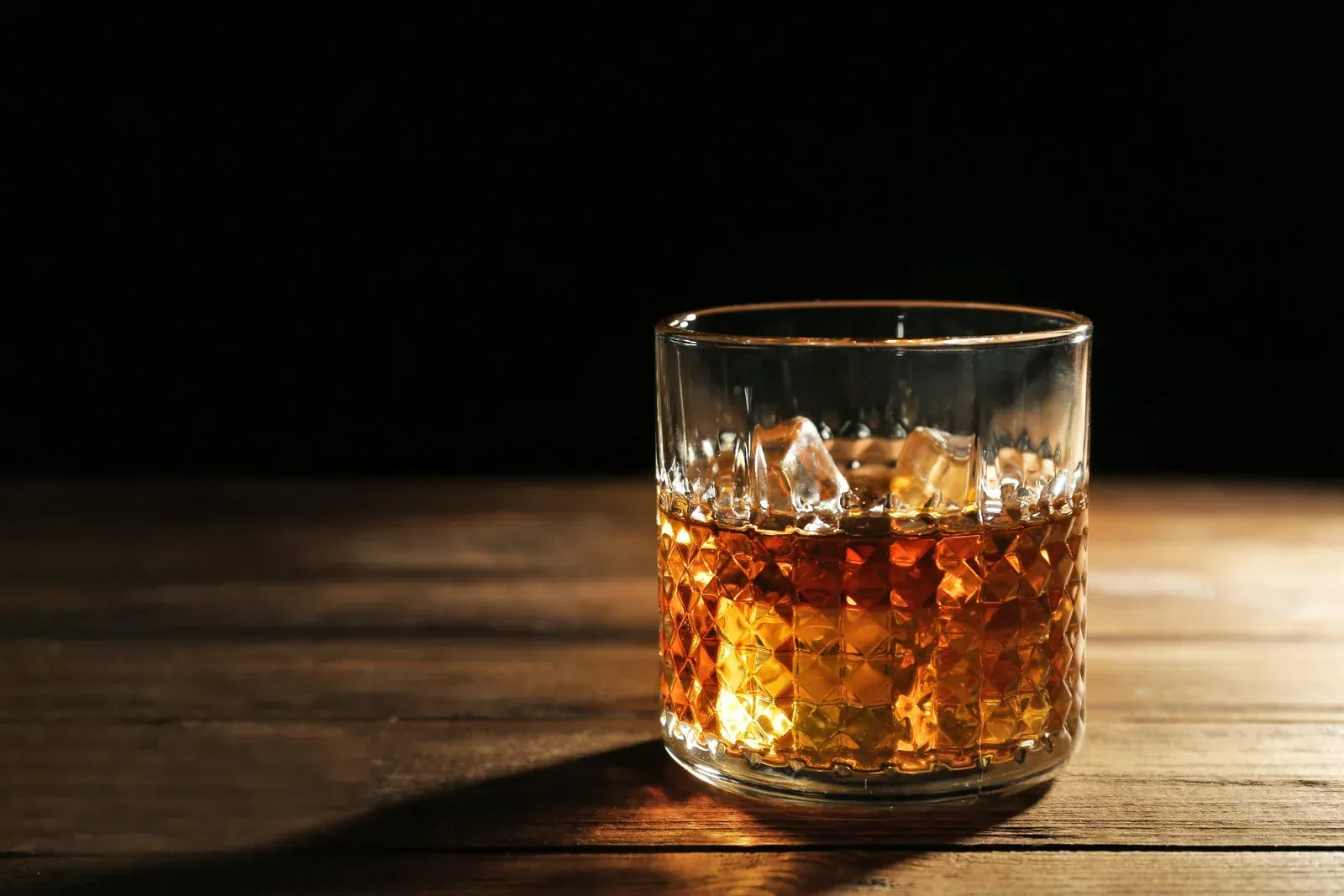 A glass of amber whiskey with ice on a wooden table against a dark background.