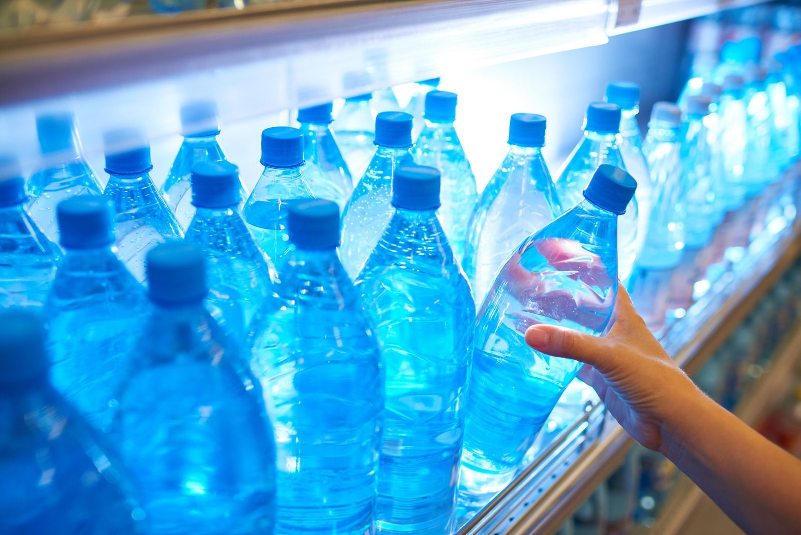 Hand reaching for blue plastic water bottles in a refrigerated store aisle