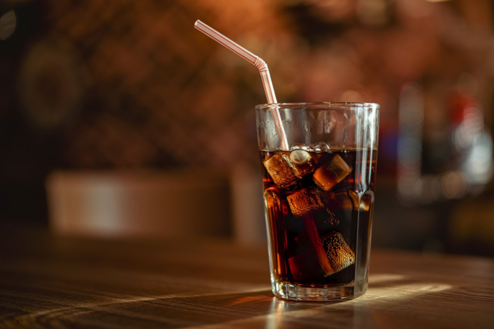 Glass of cola with ice and striped straw on a wooden table in a dim, warm-lit setting