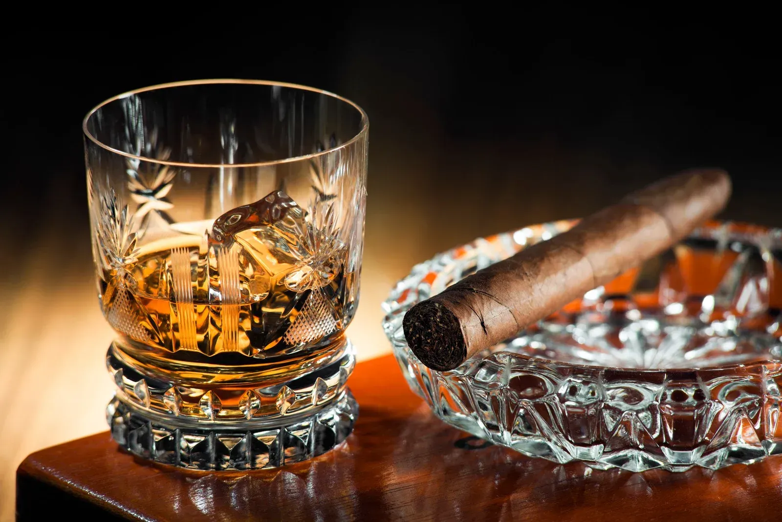 Whiskey in a glass beside a cigar on a cut-glass ashtray on a wooden table