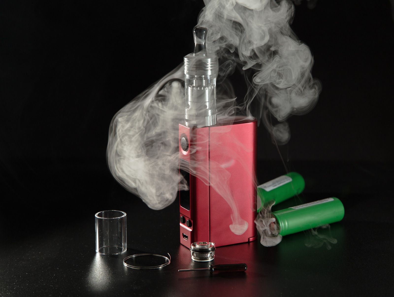 Pink vape device on black surface with billowing white vapor and a green battery pack nearby