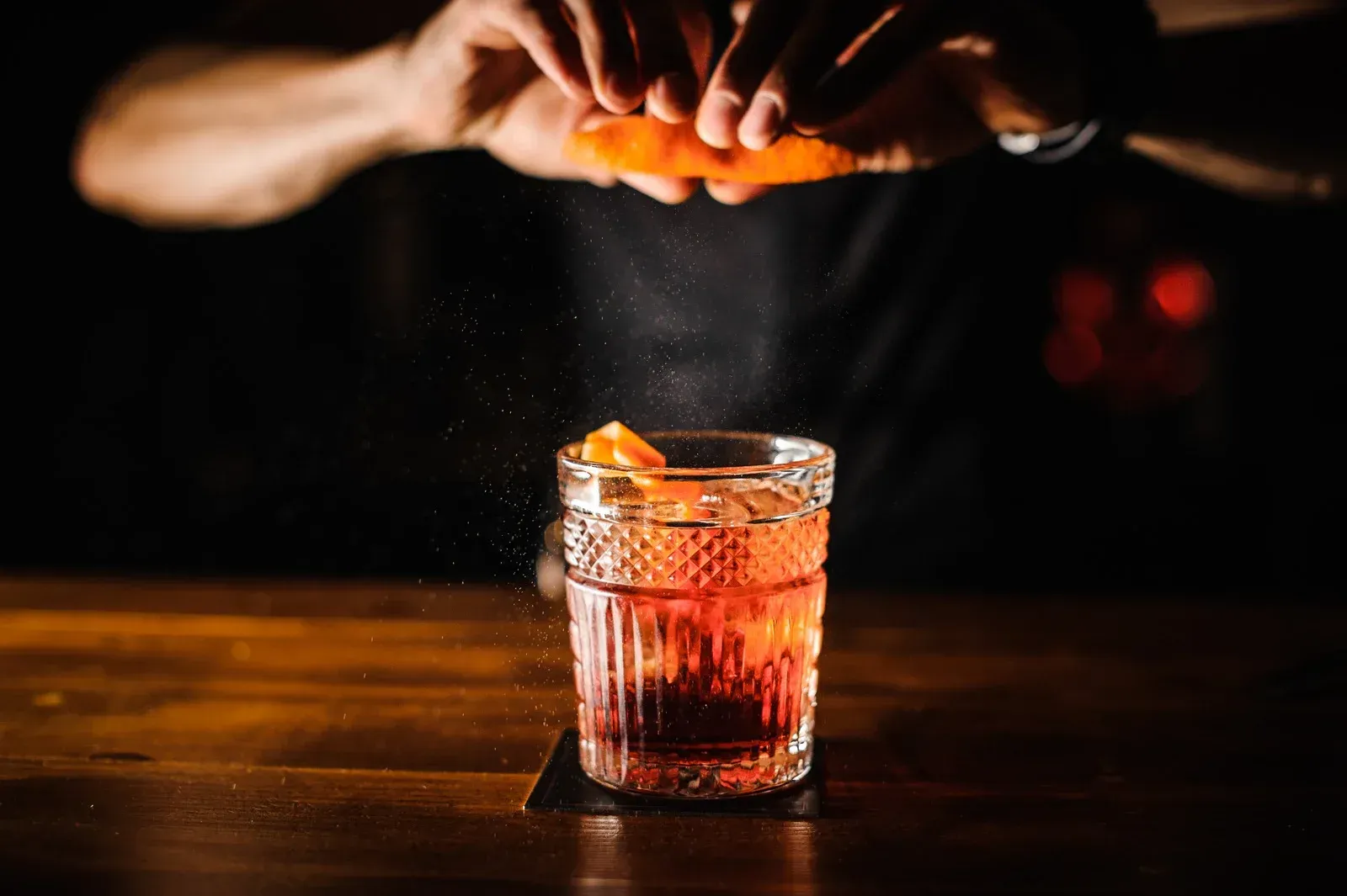 Hands squeezing an orange peel over a red cocktail in a glass on a dark bar counter.
