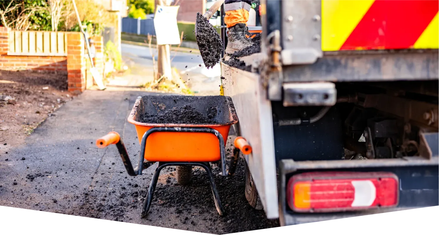 An orange wheelbarrow filled with fresh asphalt sits behind a utility truck on a paved road under construction.
