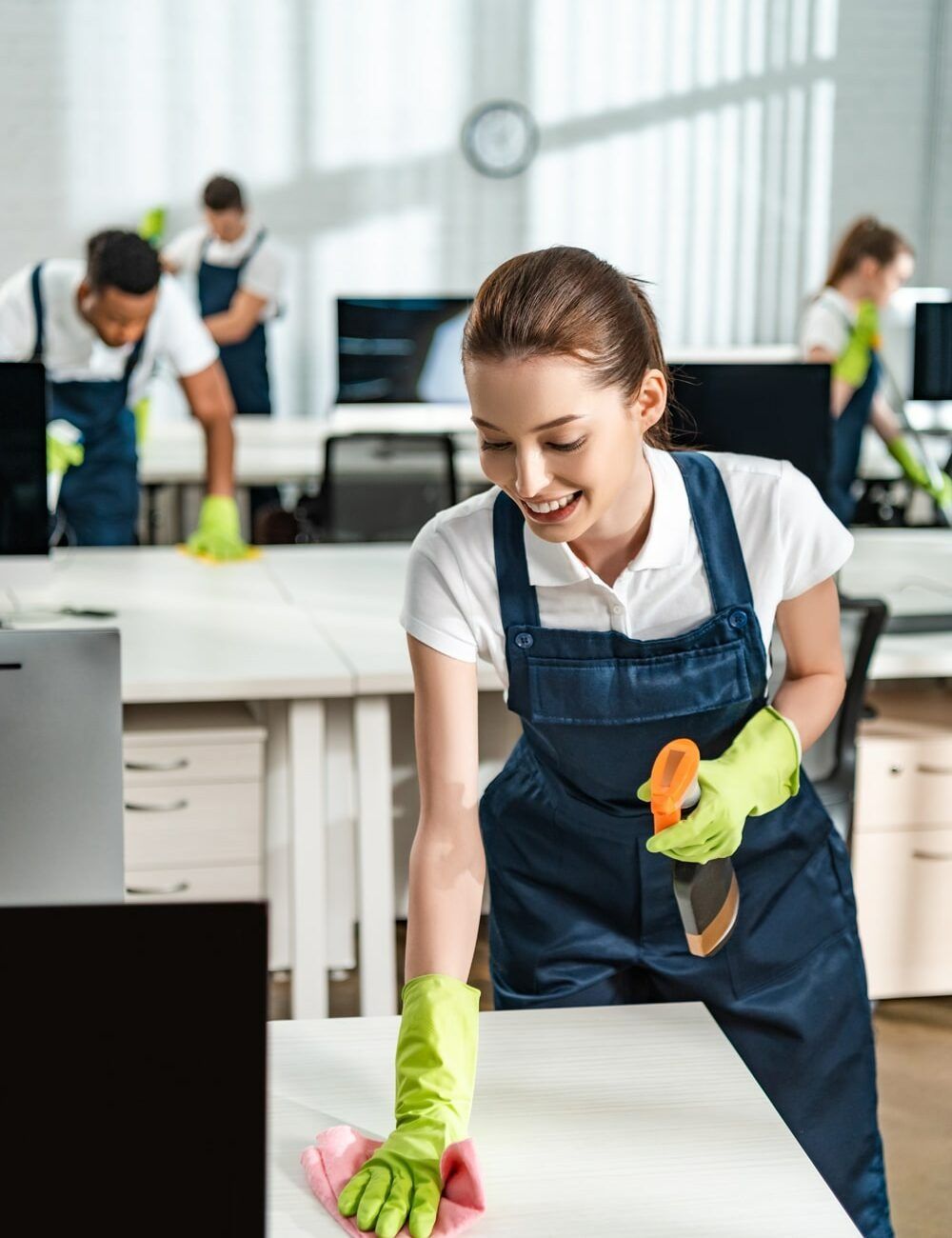 Office cleaners wiping desks, wearing green gloves and navy overalls.