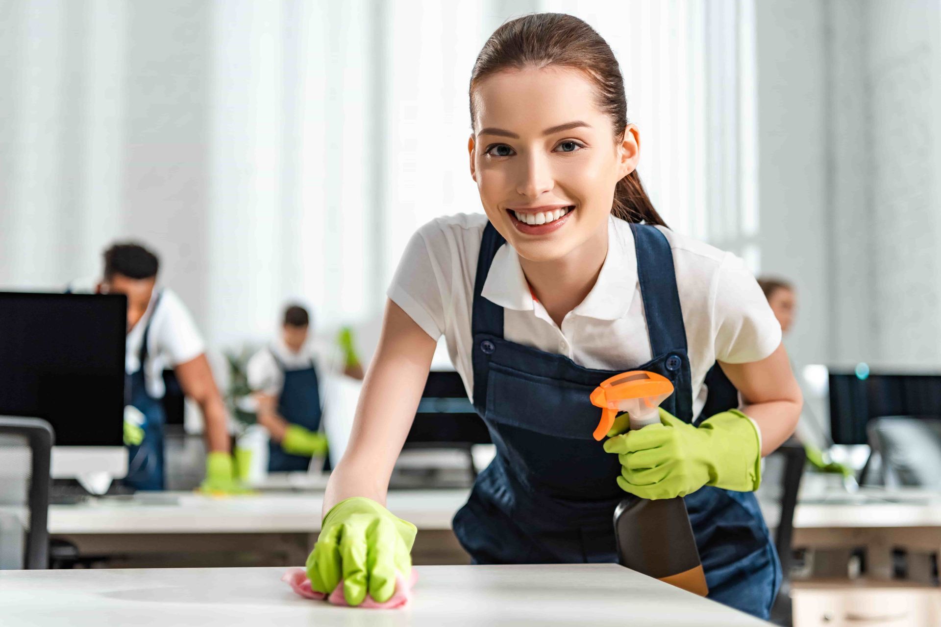 Woman in uniform smiling, cleaning office desk with spray bottle and gloves; team cleaning in background.