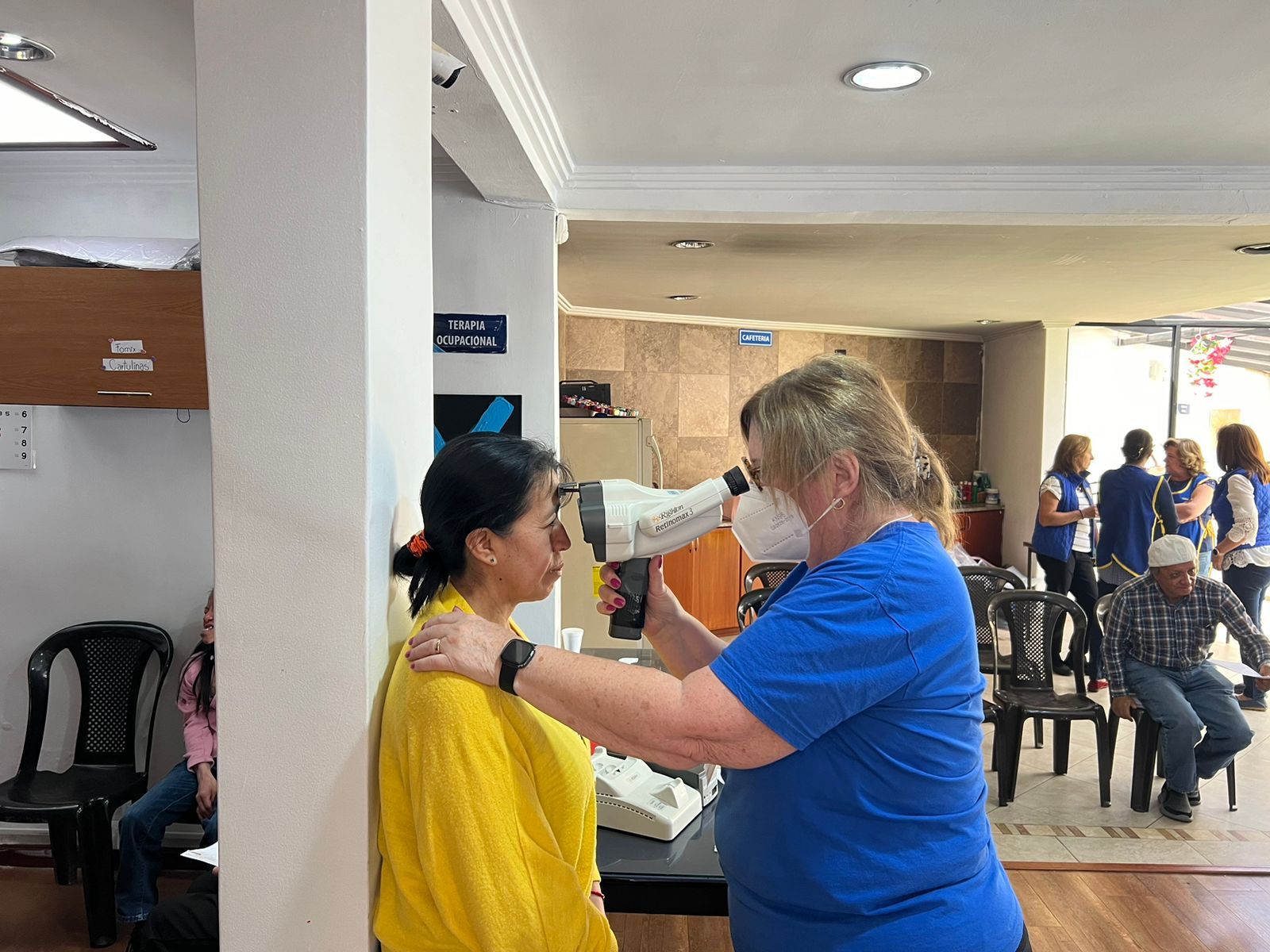 Una mujer de amarillo se somete a un examen de la vista, realizado por una persona de azul, en una clínica. 