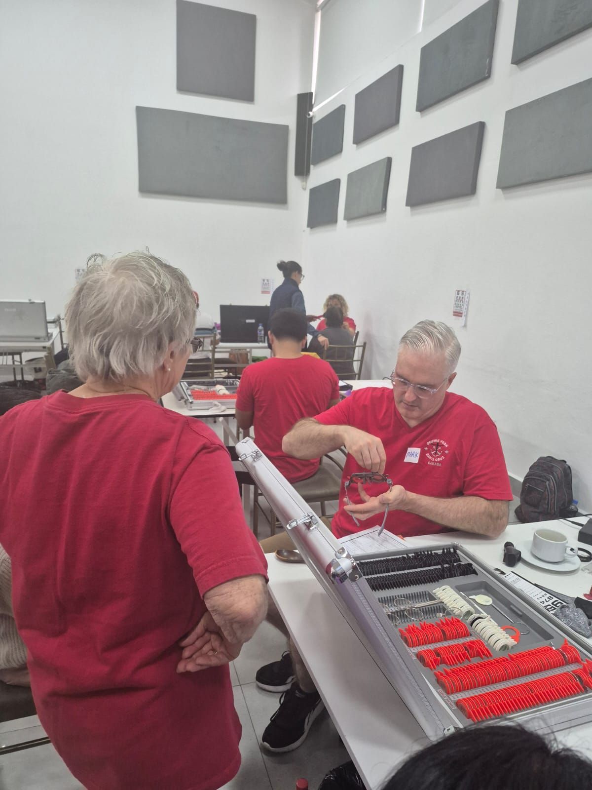 Dos personas con camisas rojas ensamblan un dispositivo en una mesa. Otros están sentados en una sala bien iluminada.