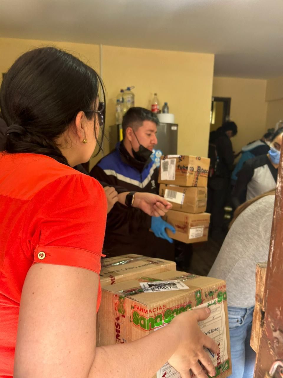 Personas repartiendo cajas de suministros en interiores. Una mujer con camisa roja lleva cajas