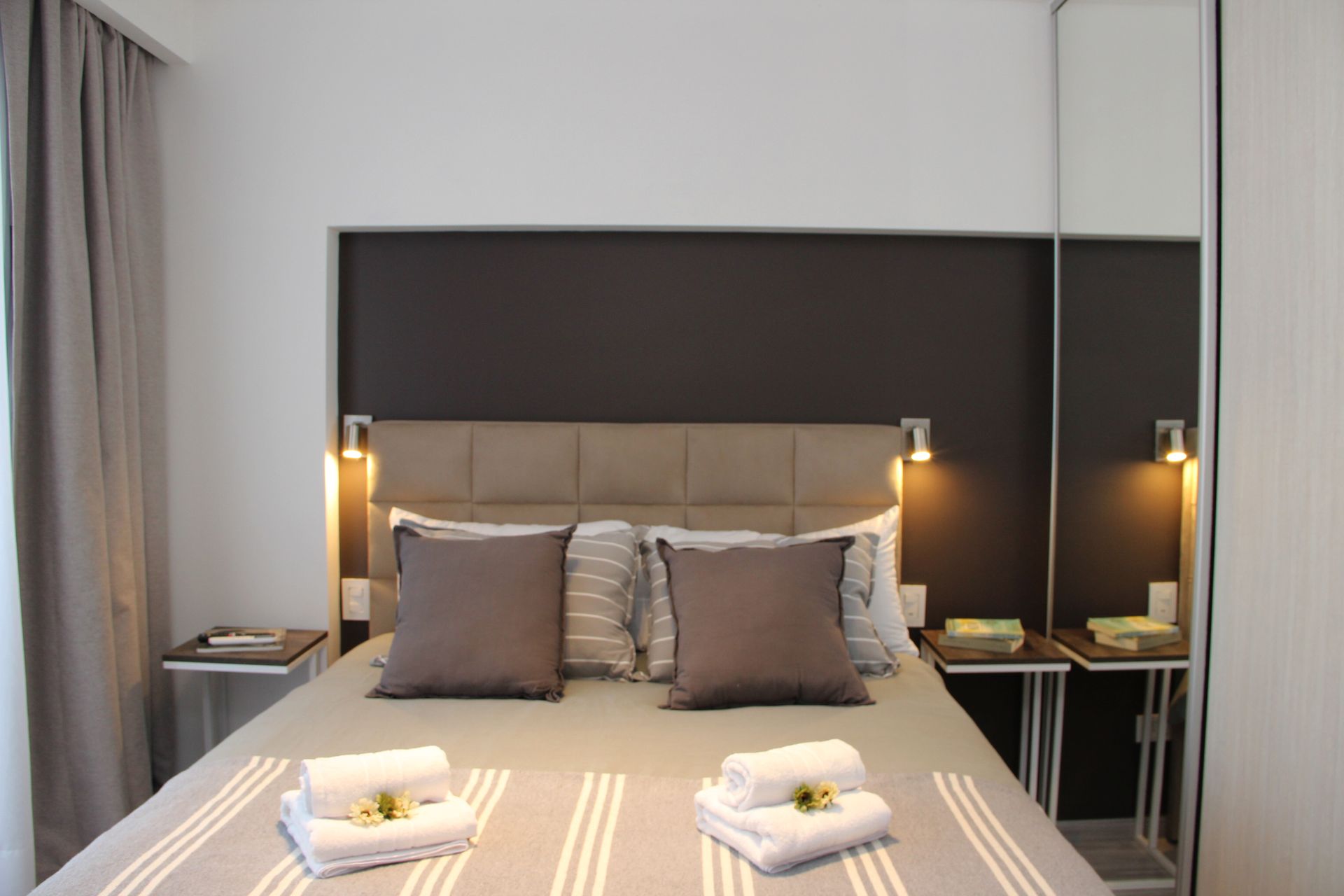 Modern bedroom with a neatly made bed, gray pillows, bedside lamps, and folded towels at the foot of the bed