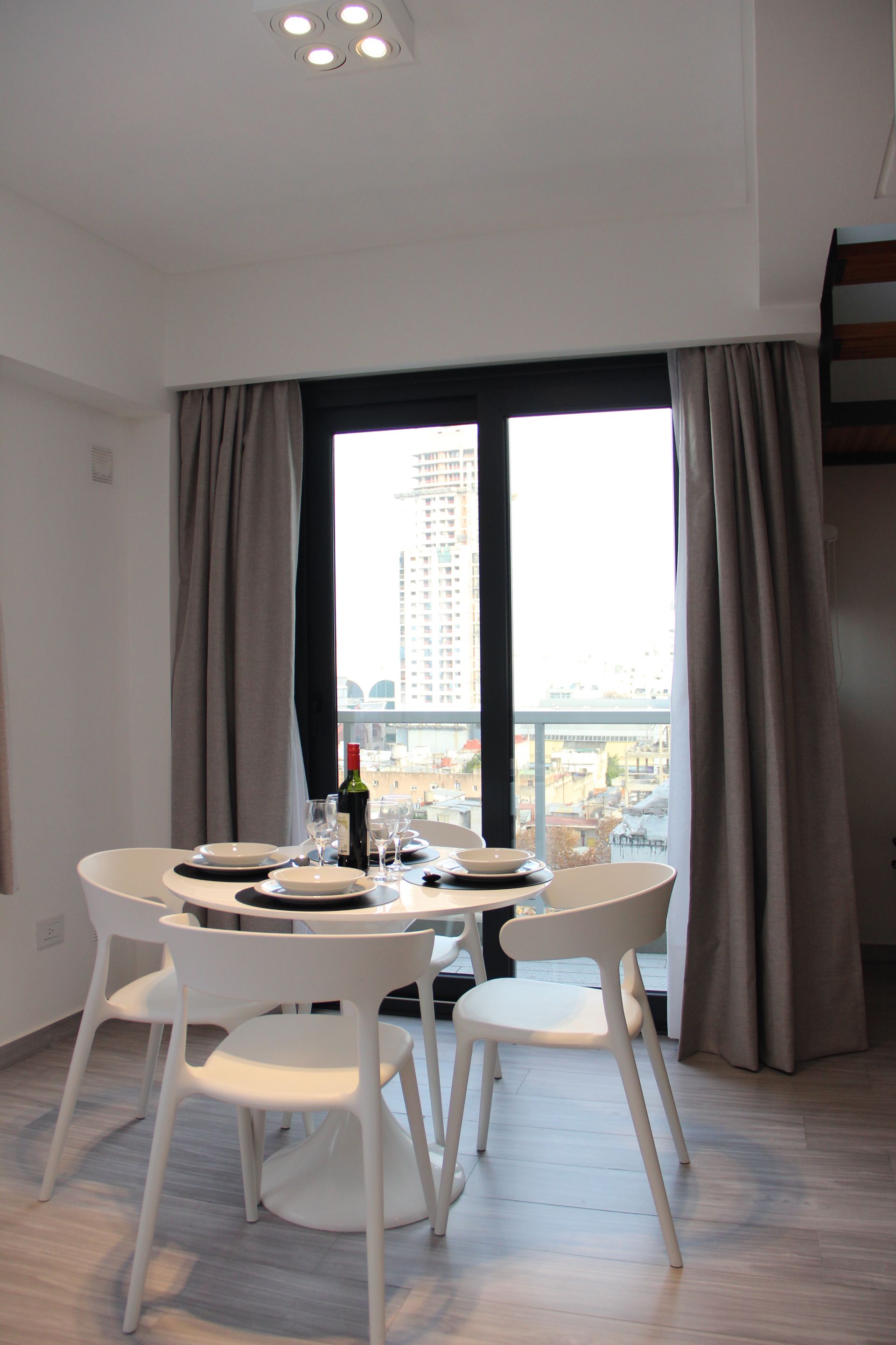 Bright dining room with round table, white chairs, curtains, and a window view of tall buildings.