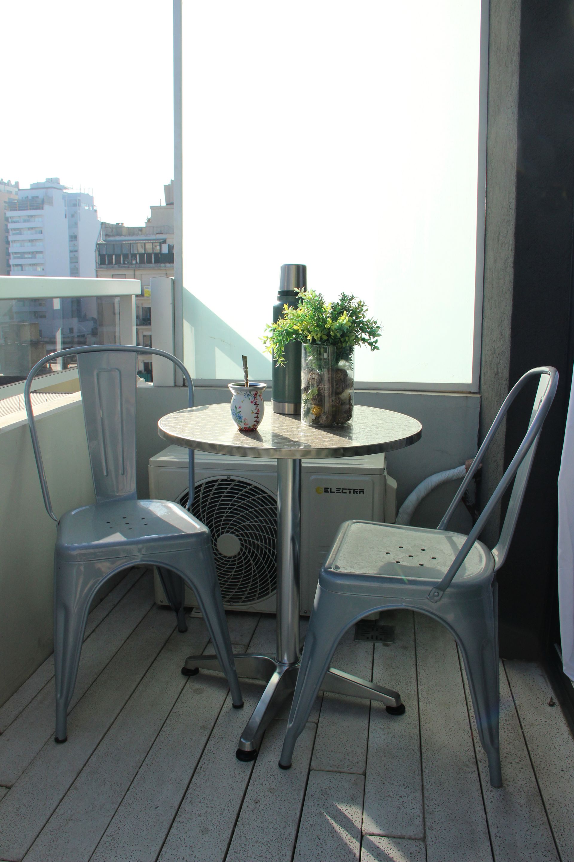 Small balcony with metal table and chairs, potted plant, and city view beyond the railing.