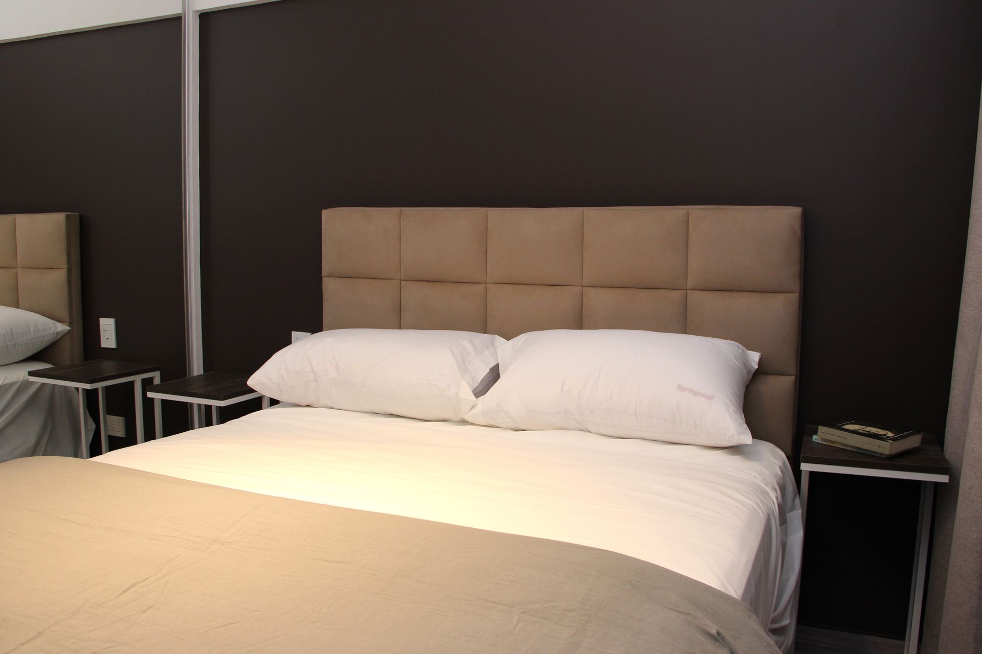 Neatly made bed with beige headboard in a dark hotel room, with a mirror and chair beside it