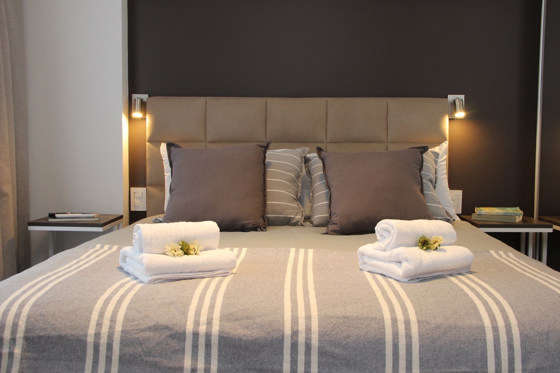 Neatly made bed with gray striped blanket, pillows, and folded towels in a modern bedroom with bedside lamps