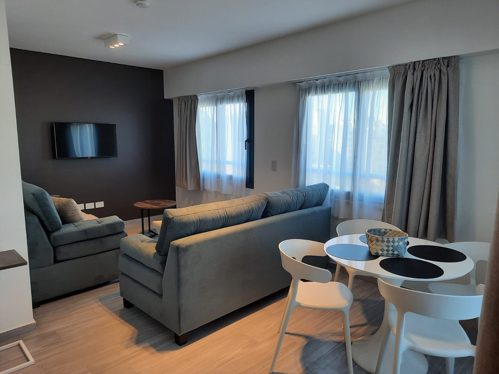 Modern living room with gray sectional, wall-mounted TV, large windows, and dining table with white chairs