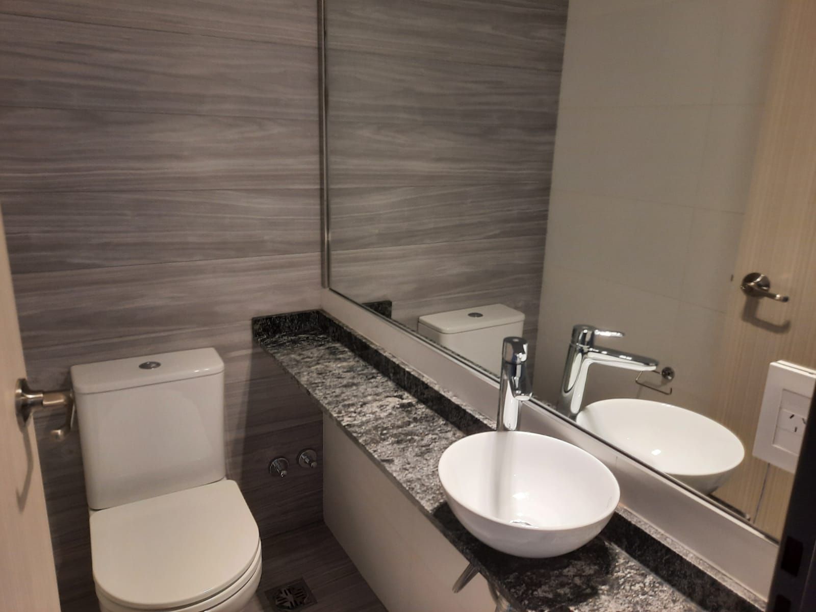 Modern restroom with toilet, granite counter, wall mirror, and two white vessel sinks