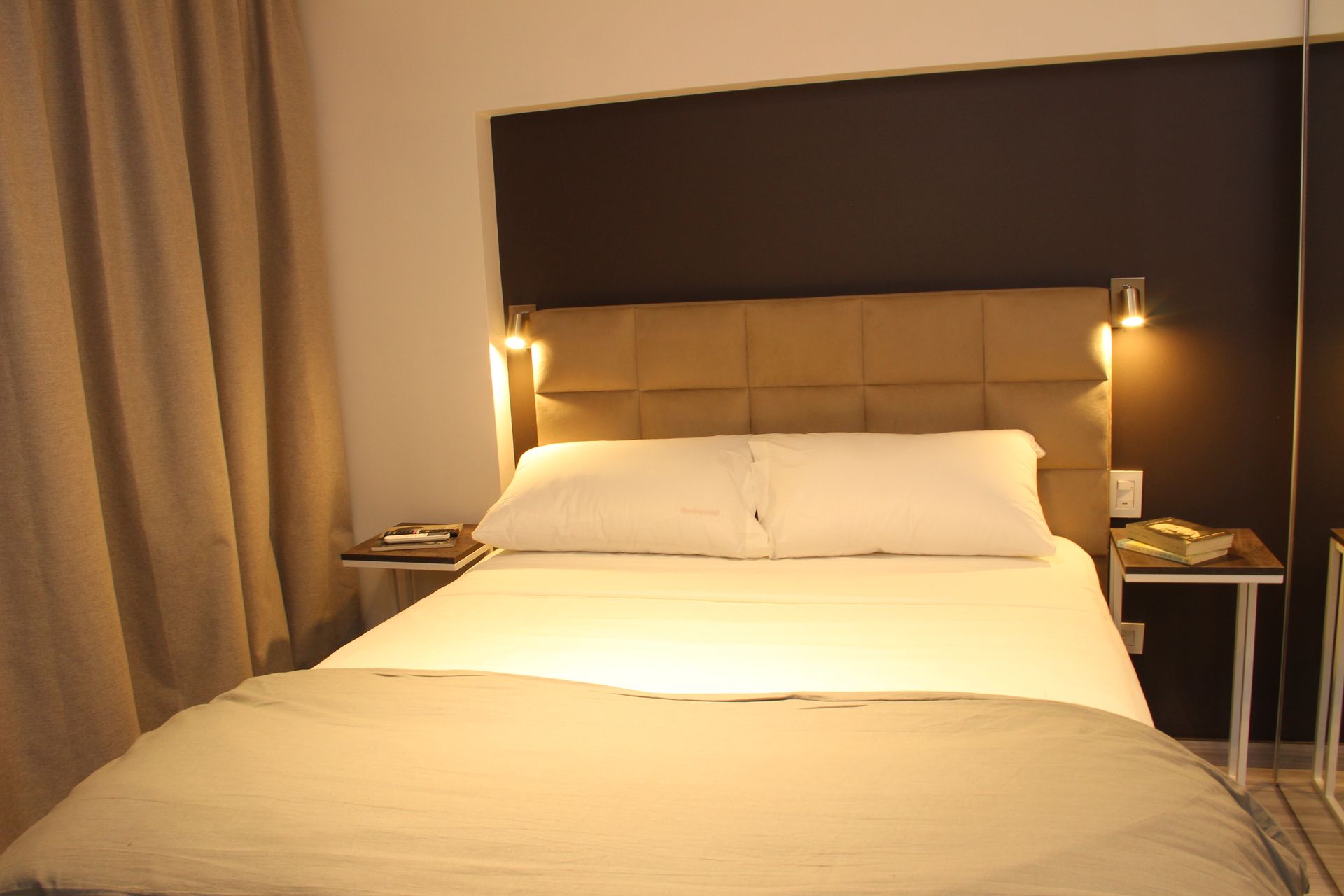 Cozy bedroom with a made bed, beige bedding, and warm bedside lamps against a dark headboard.