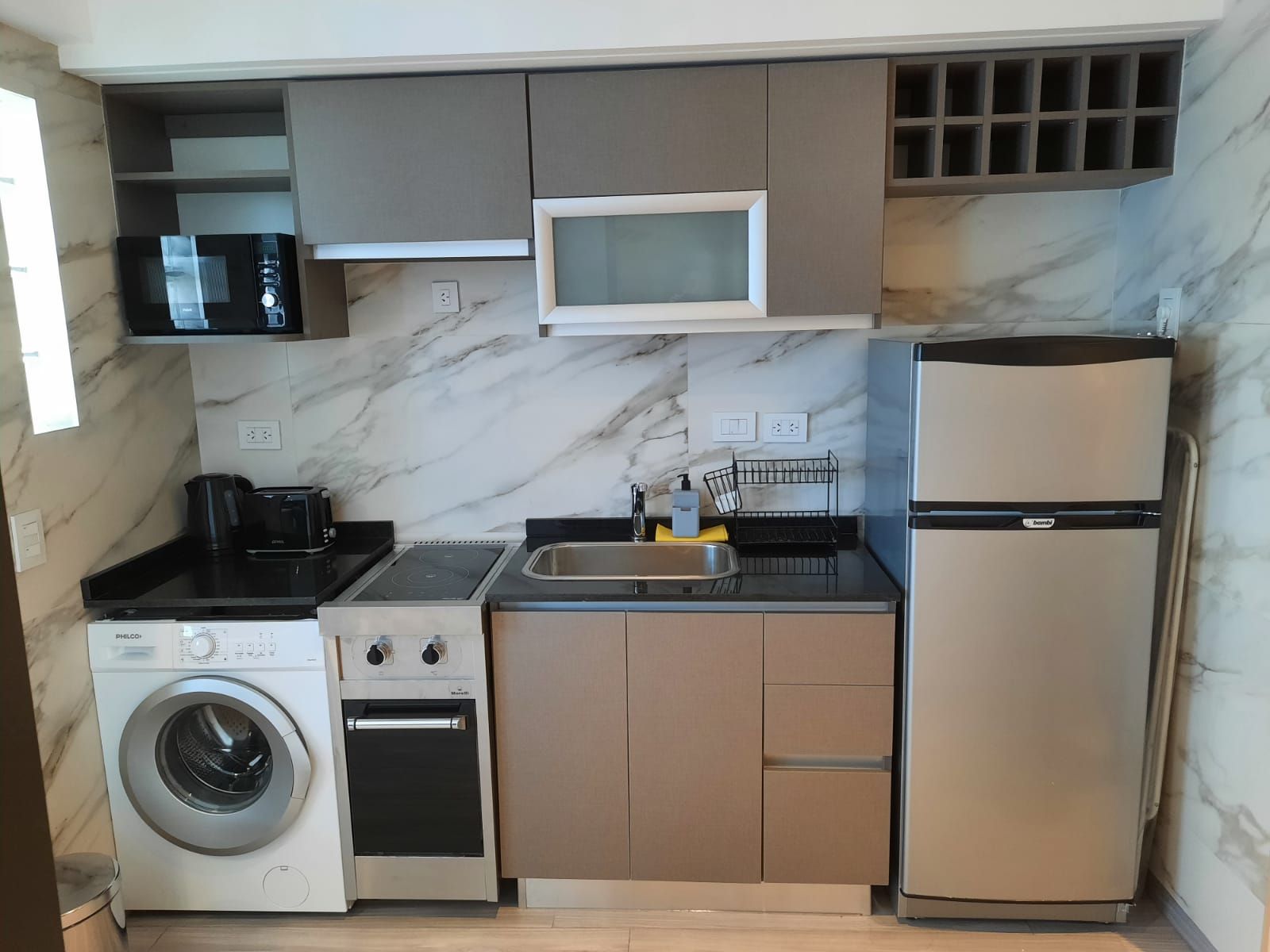 Compact kitchen with beige cabinets, marble backsplash, washing machine, stove, sink, microwave, and refrigerator.