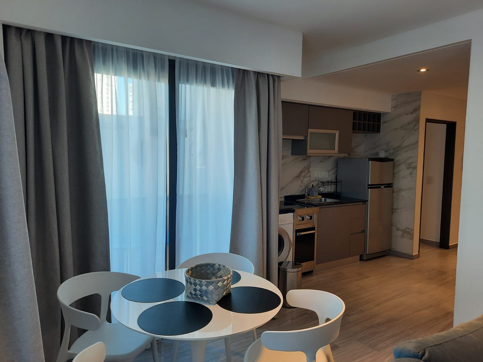 Modern apartment dining area with round table and chairs beside a compact kitchen and large window curtains