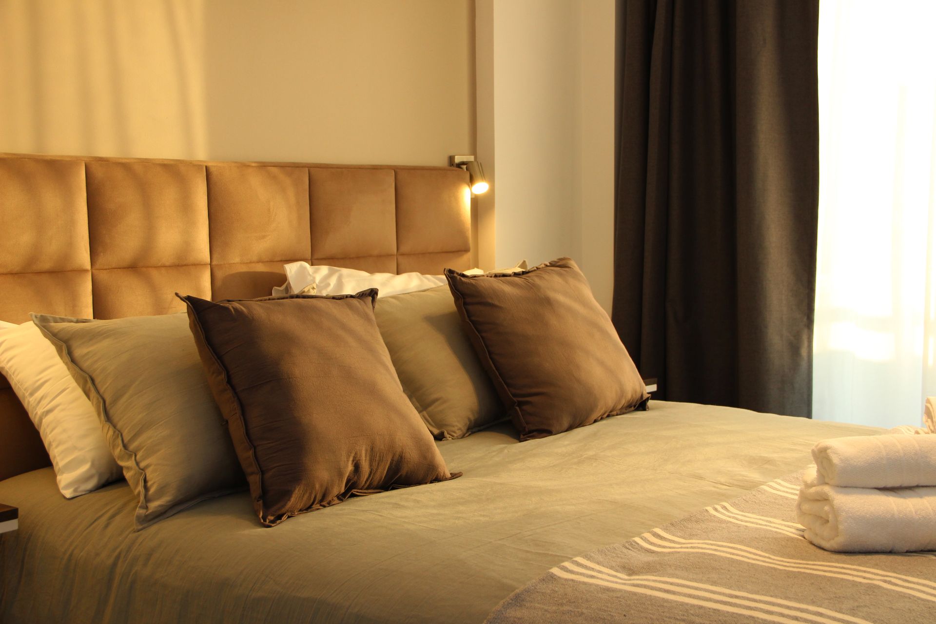 Warm bedroom with a neatly made bed, brown pillows, and soft light from a window and bedside lamp.