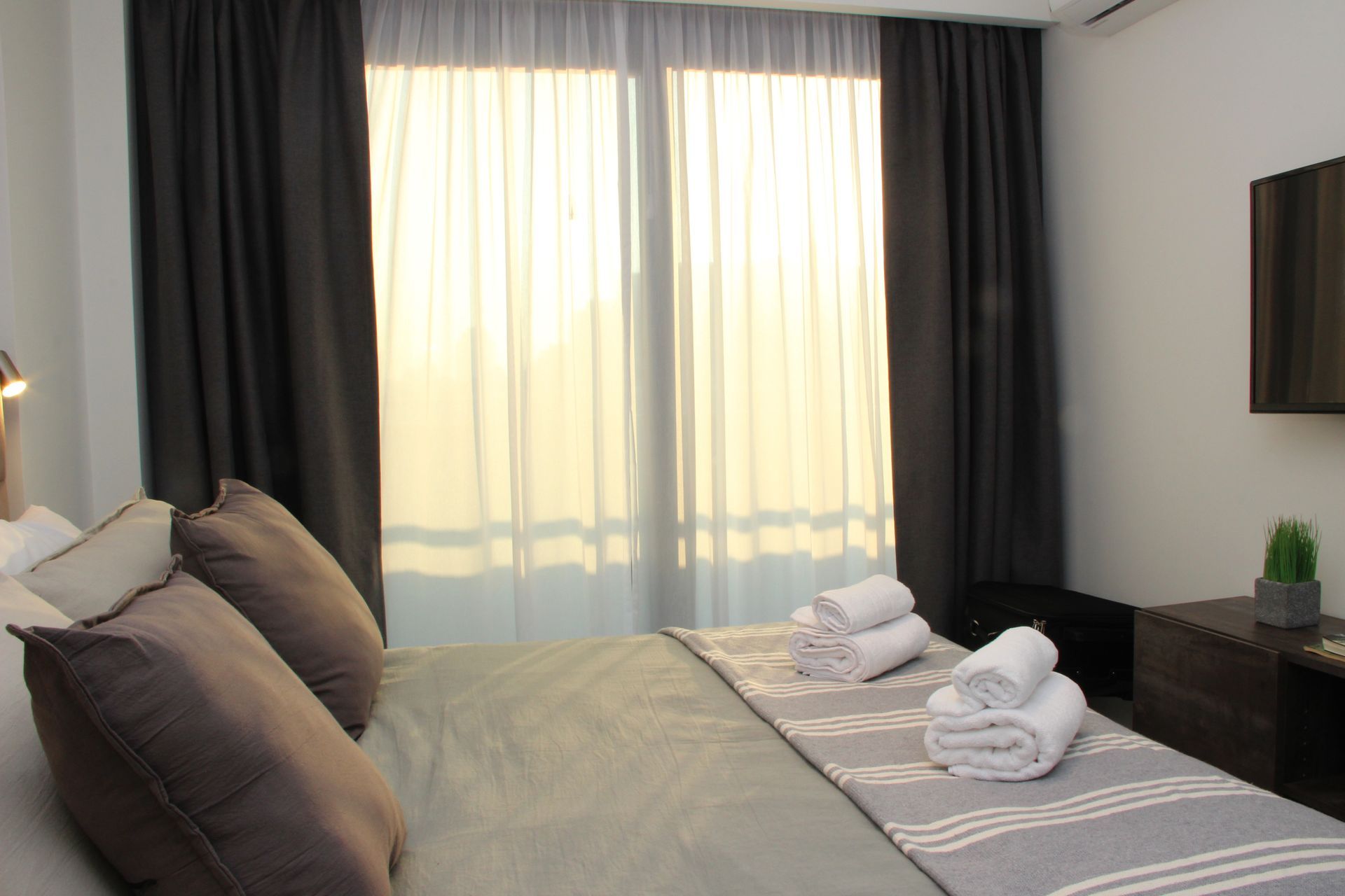 Neat hotel bedroom with a made bed, folded towels, gray curtains, and a window with sheer drapes.
