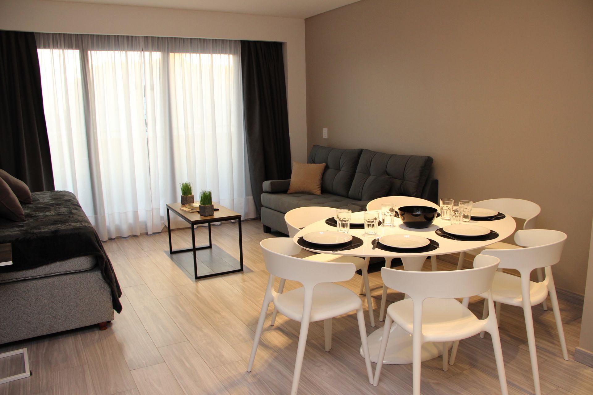 Modern dining and living room with white table and chairs, gray sofas, and large curtains by a window