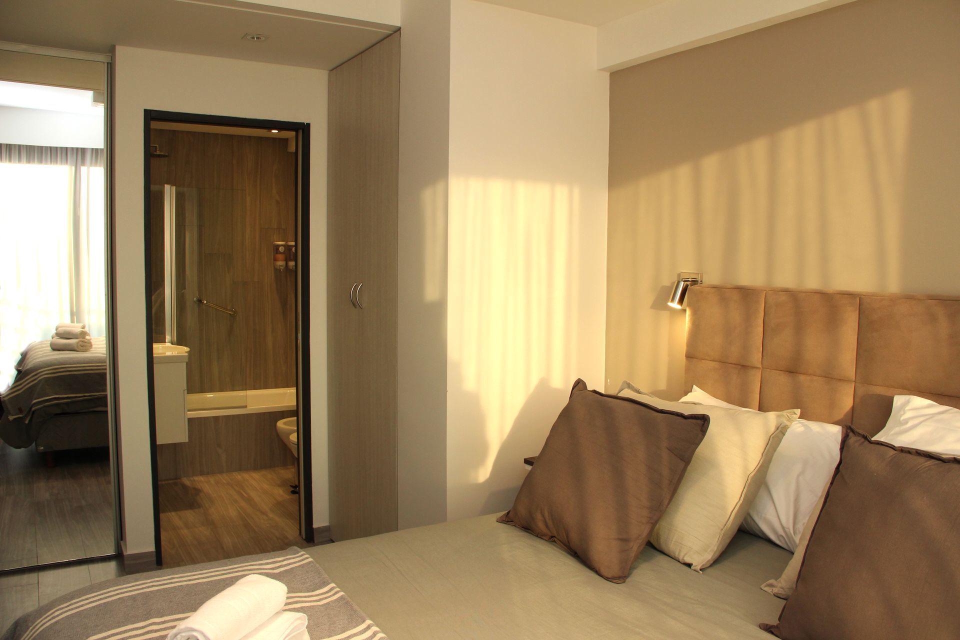 Warm hotel bedroom with beige bed, pillows, mirrored closet, and sunlight streaming through the window.
