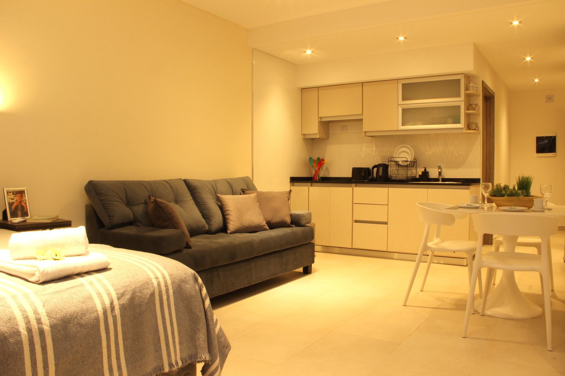 Bright modern living room with gray sofa, bed, dining table, and kitchenette under warm lighting