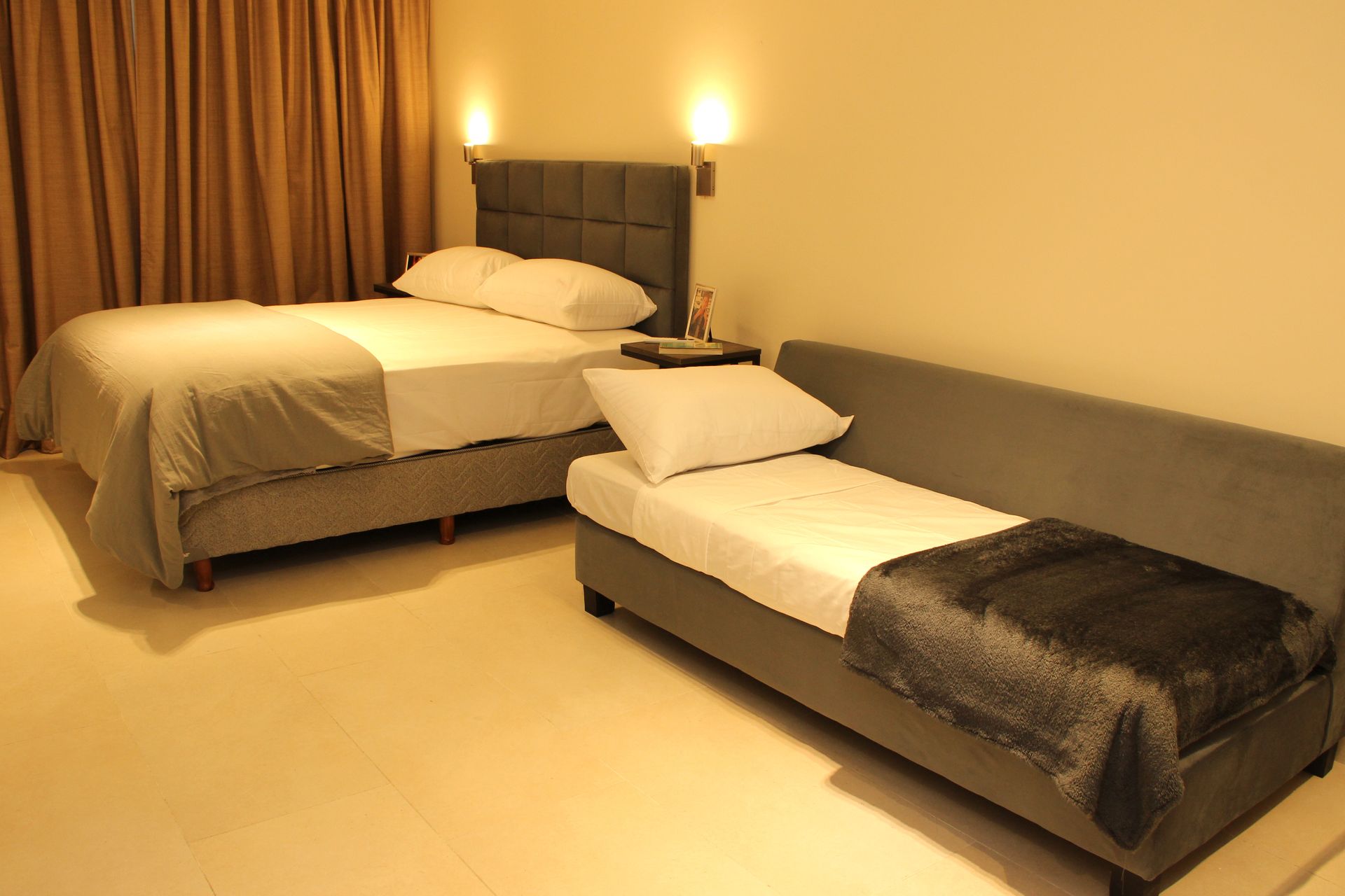 Small bedroom with a bed and two single daybeds, beige walls, and warm lamp lighting.