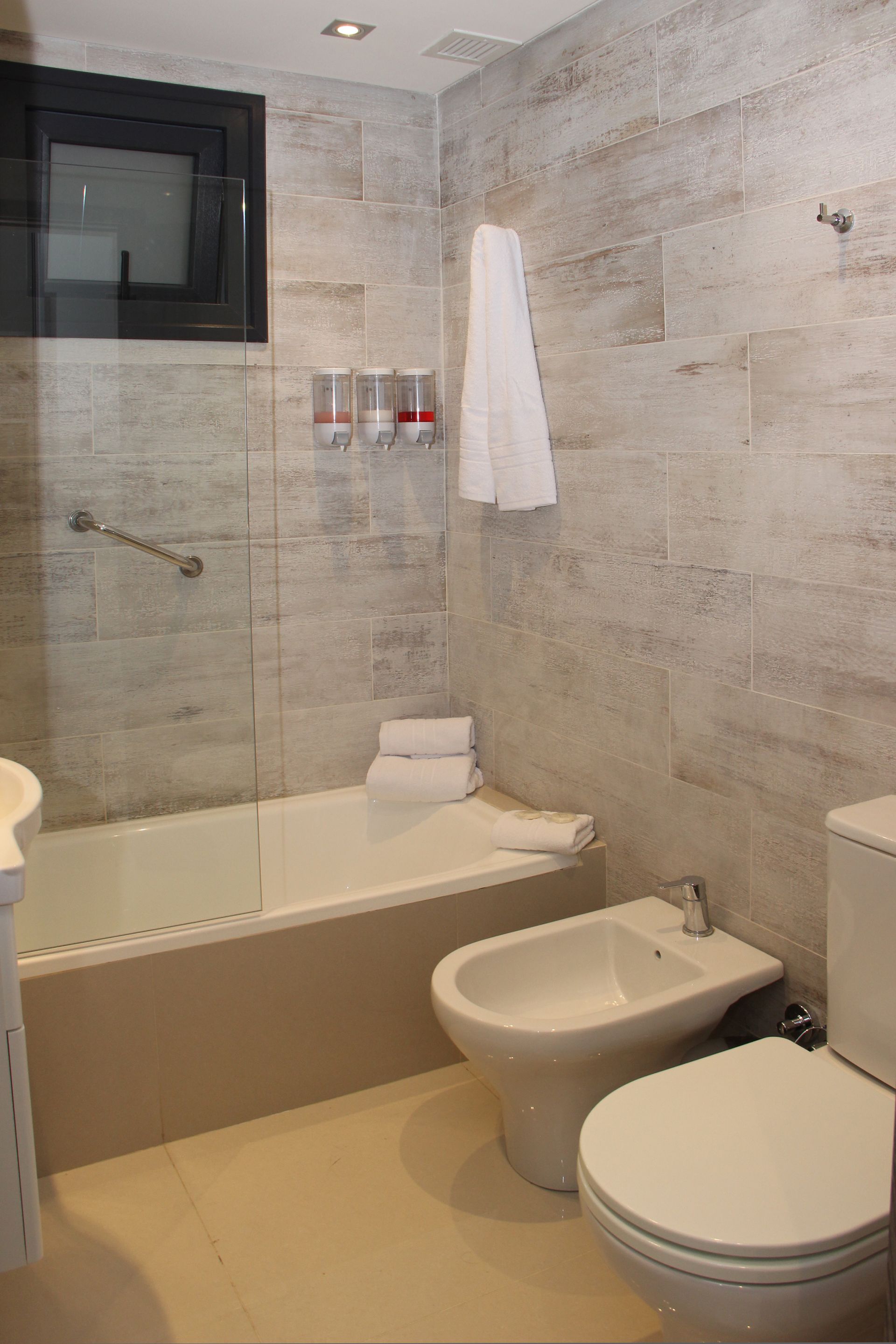 Small beige-tiled bathroom with bathtub, bidet, toilet, towel, and shower area.