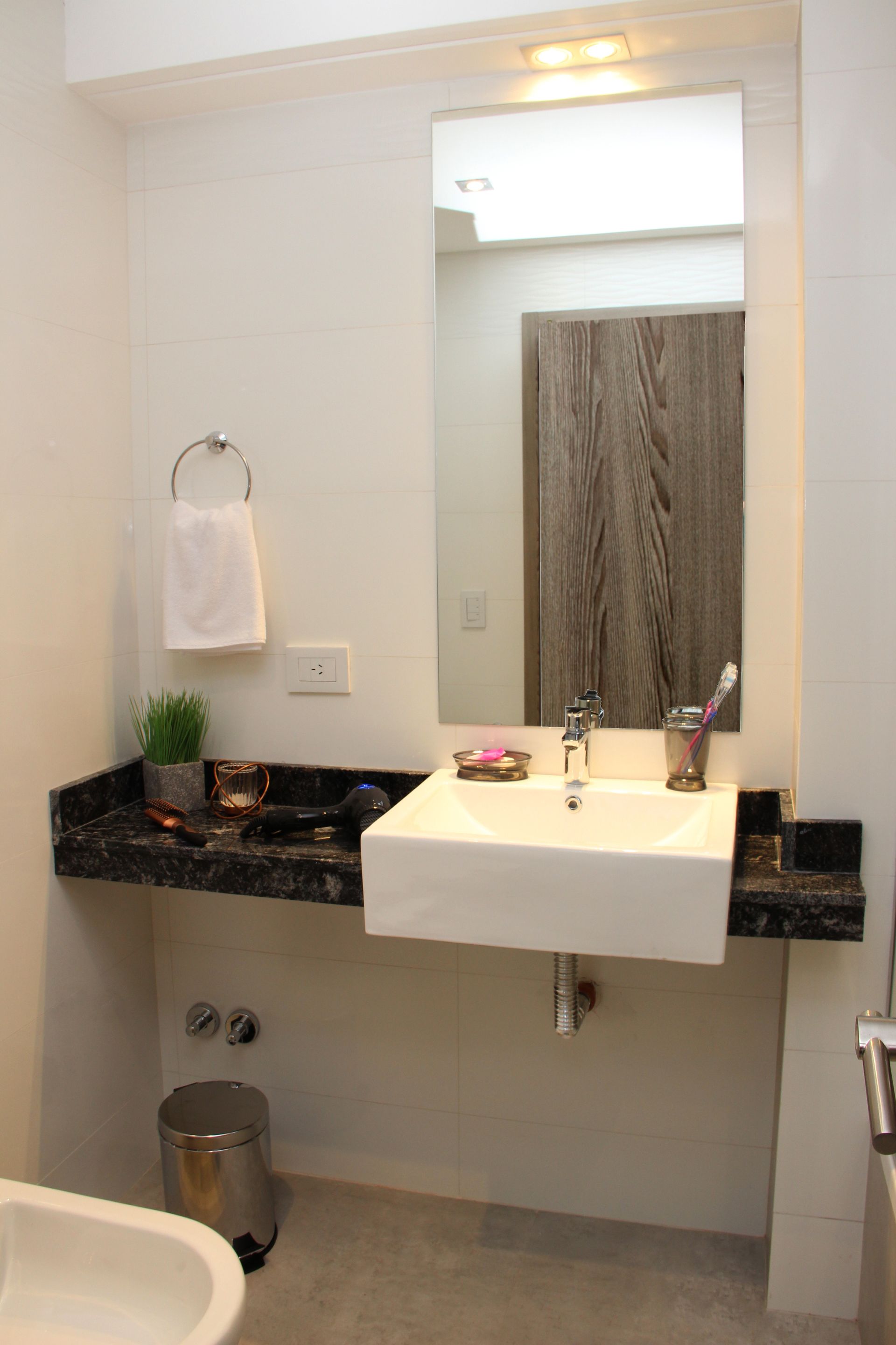 Small bathroom vanity with black countertop, white sink, mirror, towel ring, and trash can.