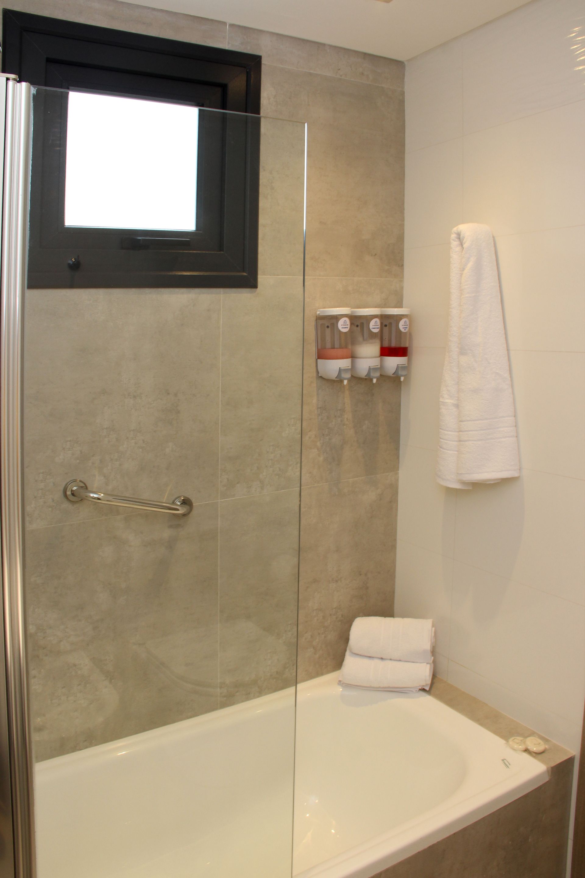 Modern bathroom with a tub, glass shower panel, wall toiletries, and a towel by a small window