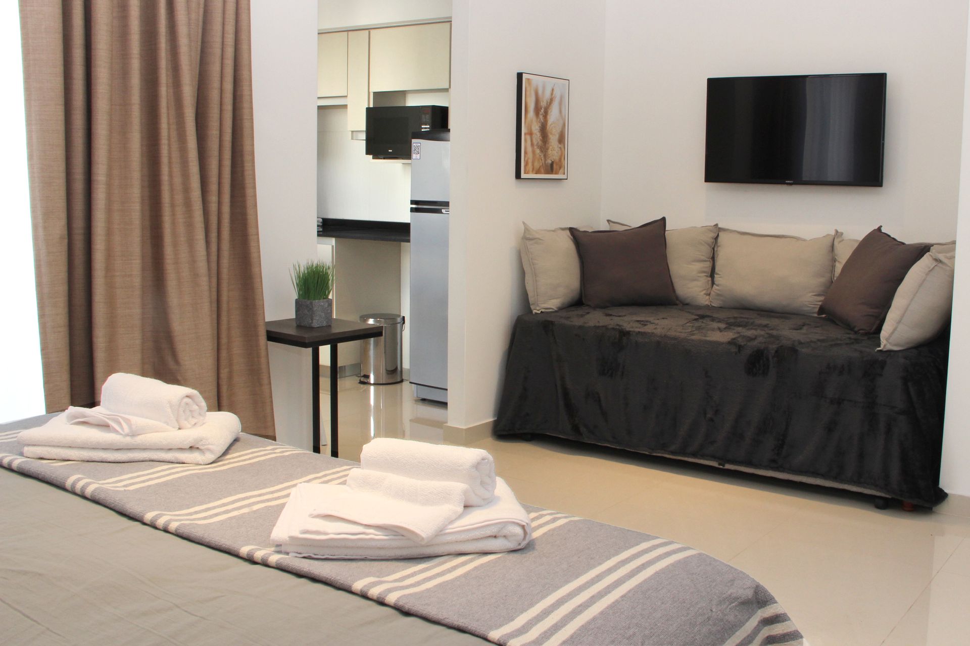 Massage room with two treatment beds, folded towels, sofa, wall-mounted TV, and kitchen visible in back