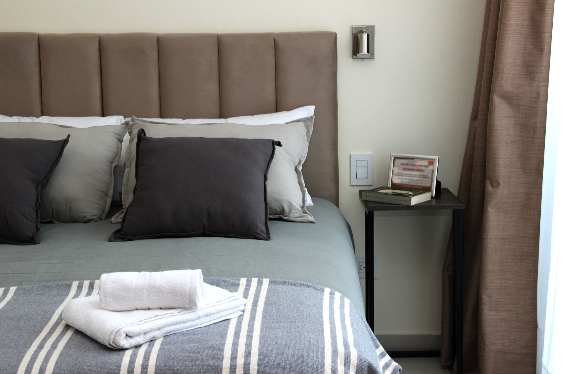 Neatly made bed with gray bedding, folded towels, and a bedside table beside a window