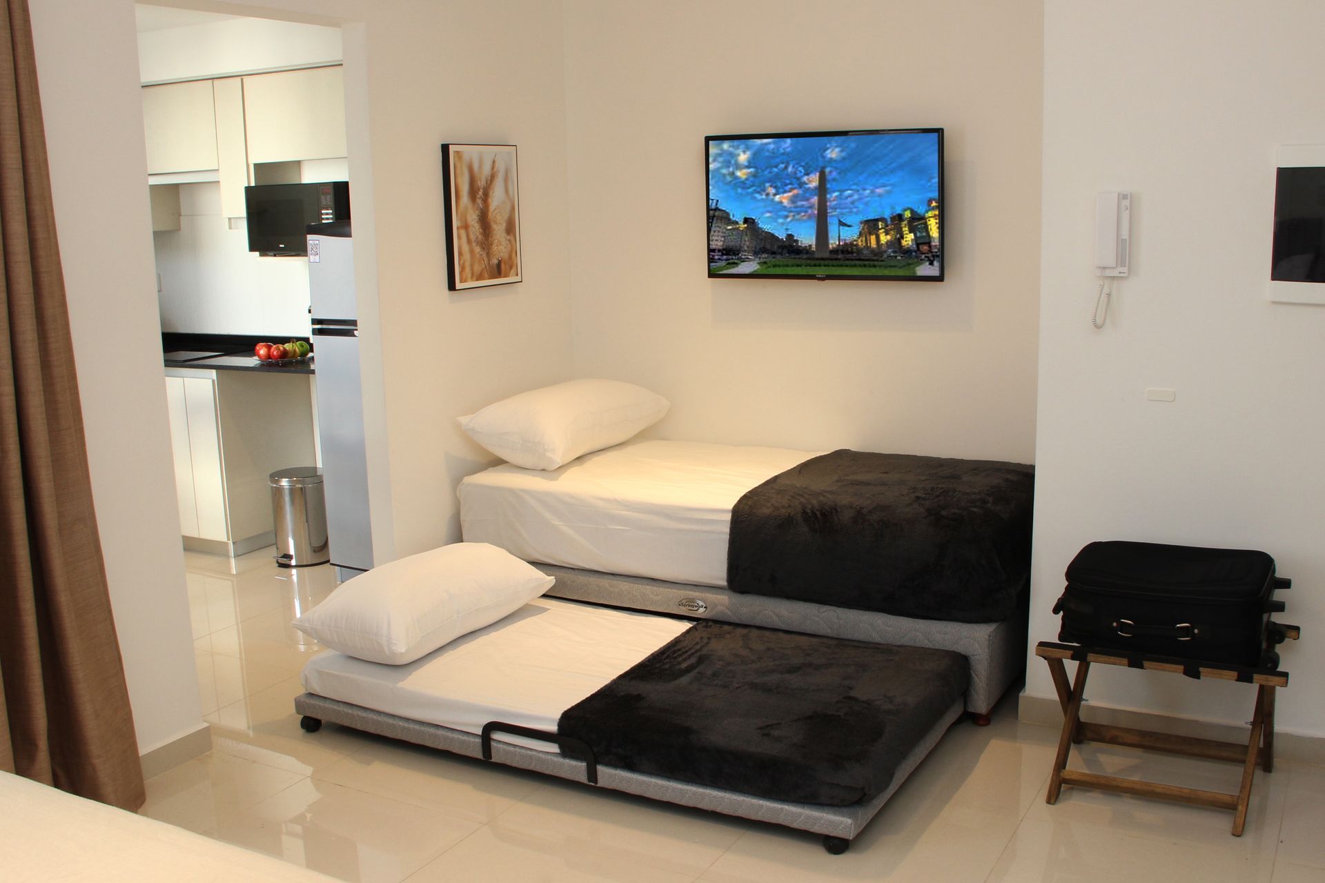 Small minimalist room with two low beds, wall-mounted TV, and an open doorway to a kitchen.
