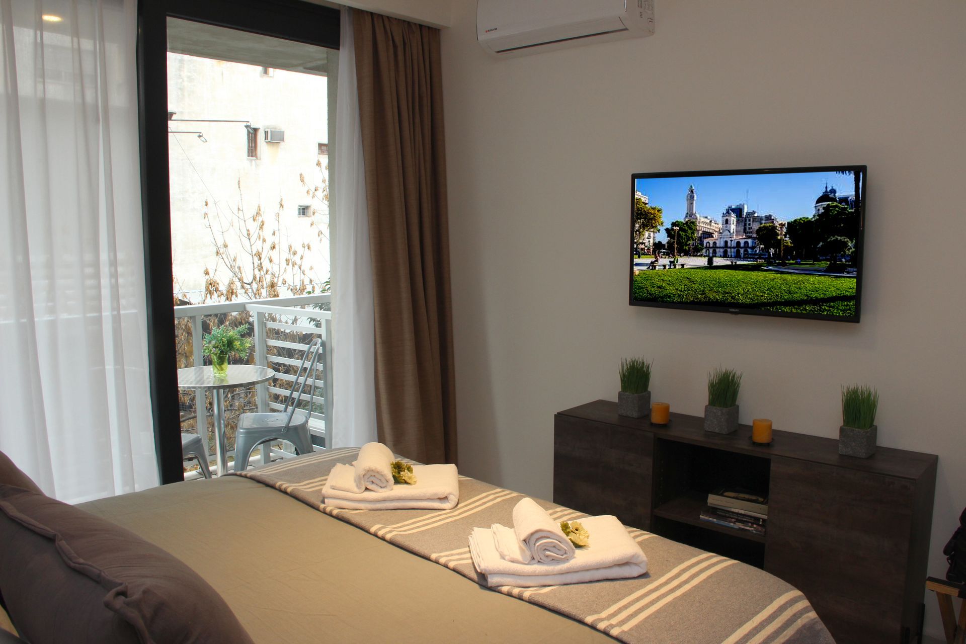 Bedroom with neatly made bed, wall-mounted TV, and a balcony door with curtains open