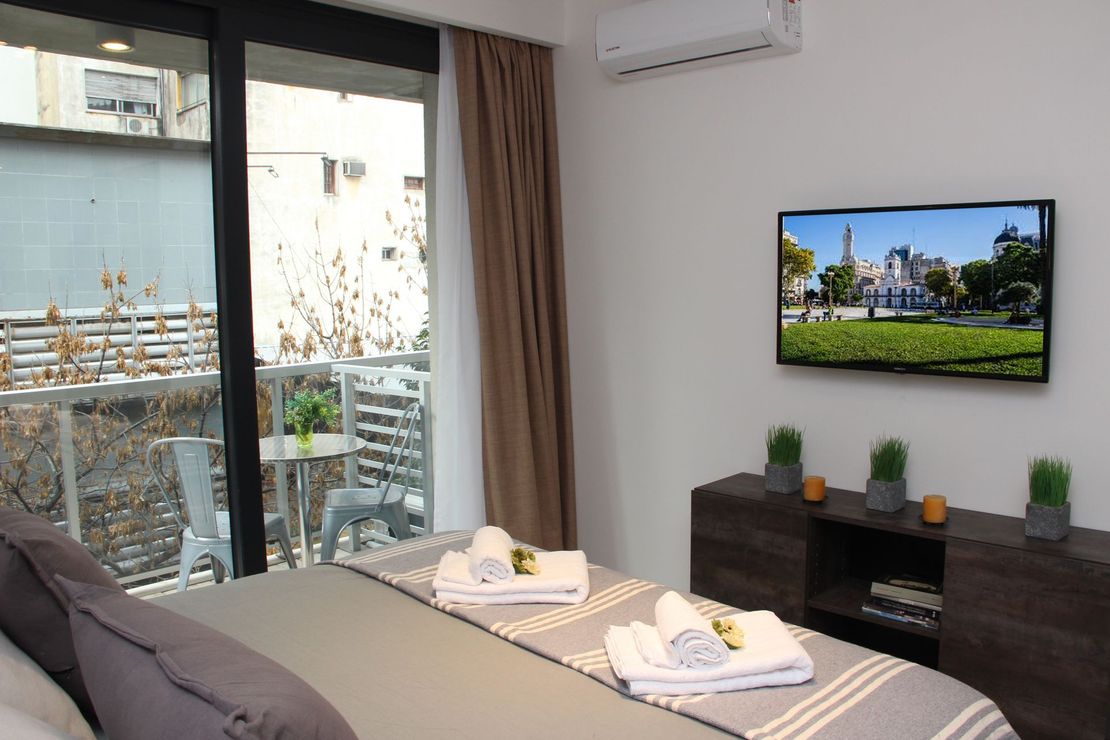 Modern bedroom with a neatly made bed, wall-mounted TV, and sliding glass door to a patio.