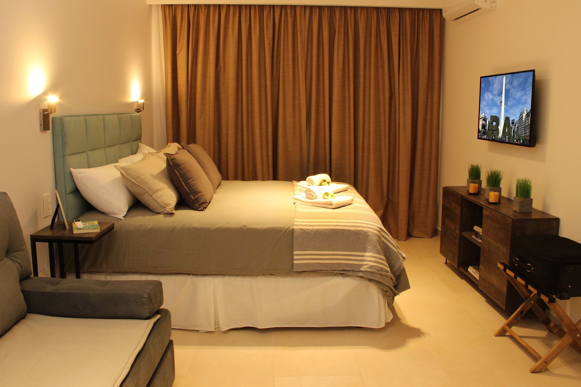 Cozy bedroom with a bed, warm lighting, curtains, wall-mounted TV, and a small seating area