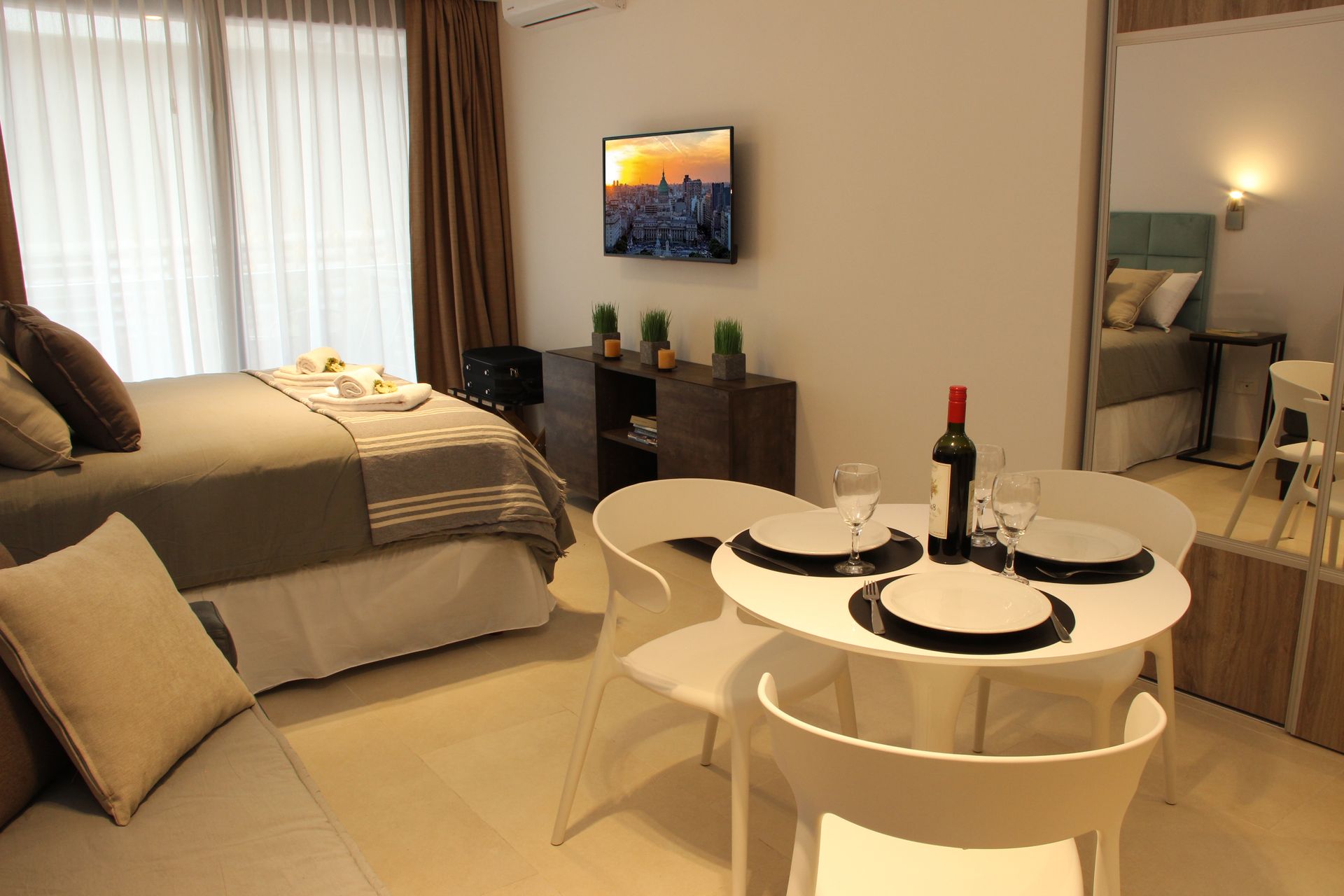 Cozy studio bedroom with a bed, dining table set for two, TV, and mirror-lit dressing area