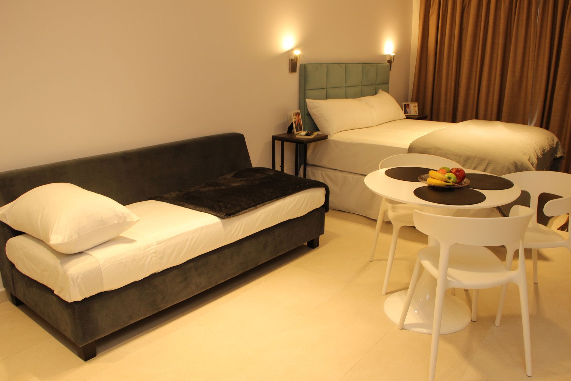 Cozy hotel room with a bed, dark sofa, and round table with fruit under warm lighting