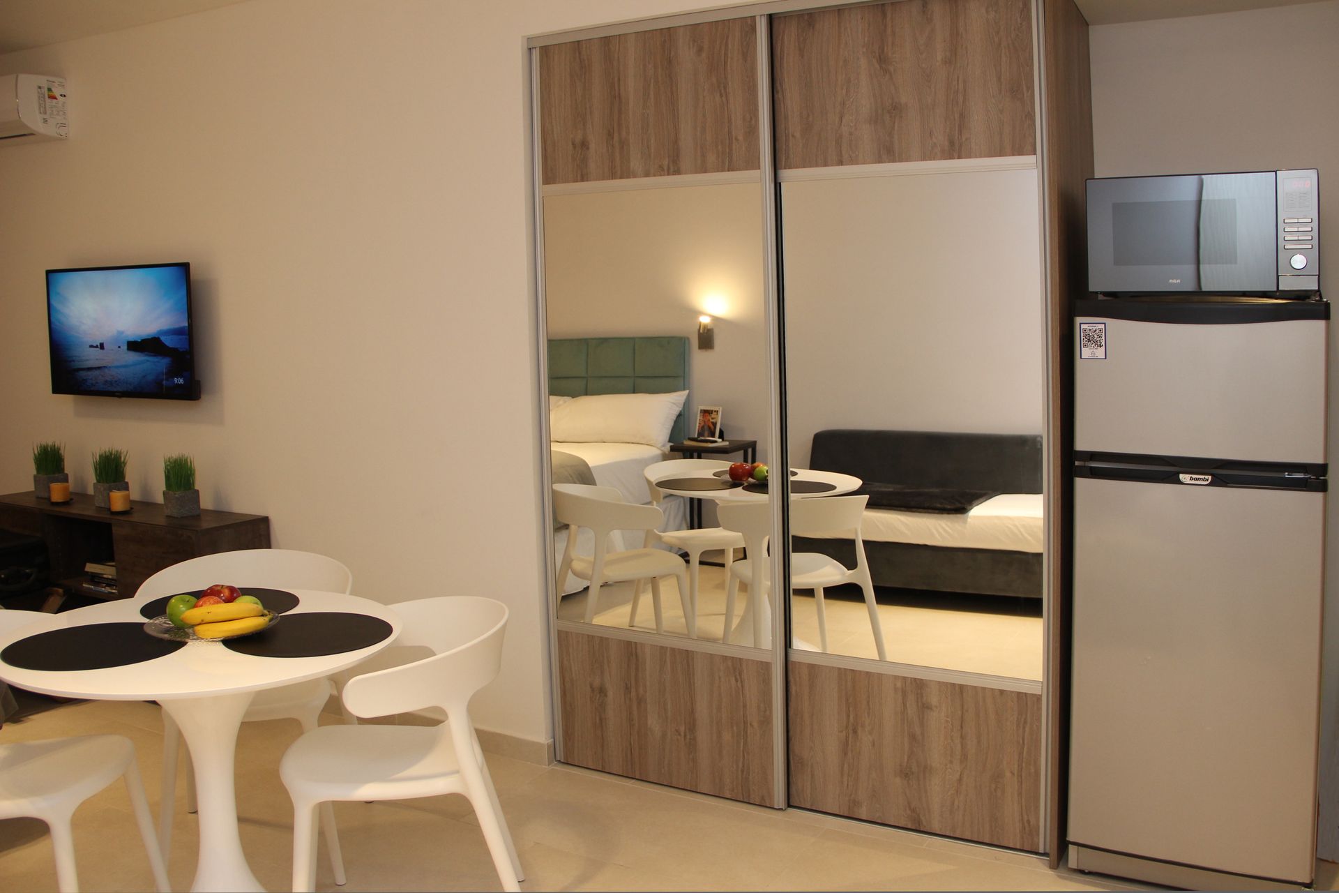 Small studio apartment with a dining table, wardrobe mirrors, bed, wall TV, and mini fridge.
