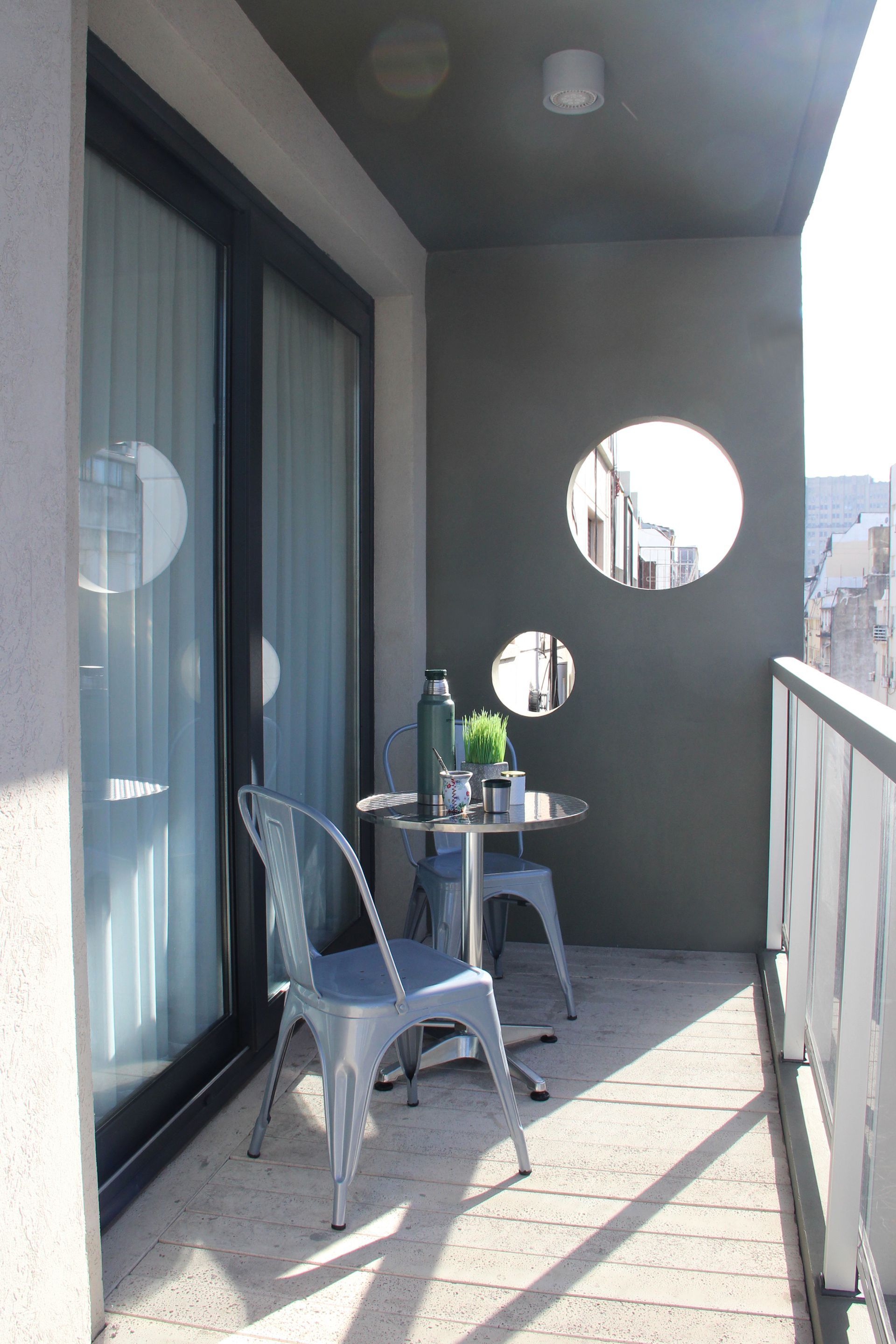 Small balcony with sliding glass doors, white chairs, a table, and circular wall mirrors in sunlight