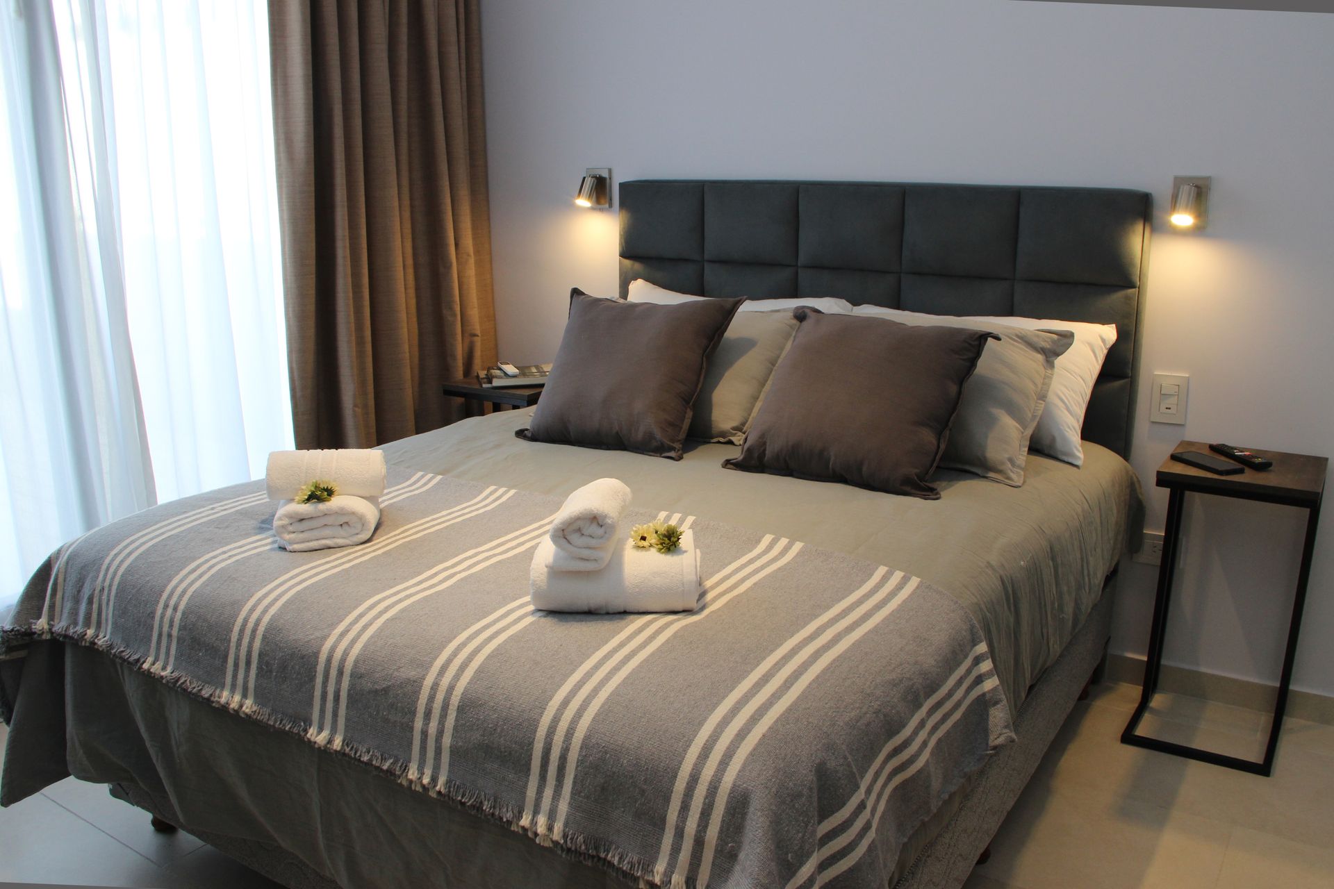 Modern bedroom with gray bedspread, dark pillows, bedside lamps, and a window with curtains