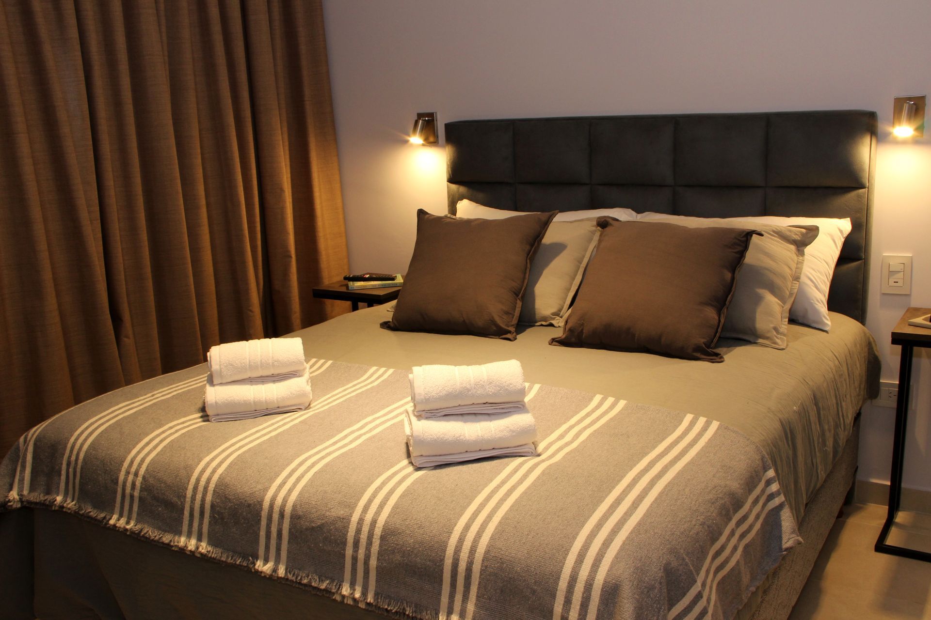 Neatly made bed with striped gray bedding, brown pillows, and folded towels in a softly lit bedroom