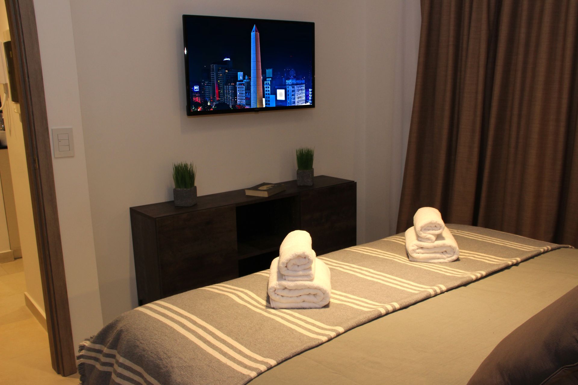 Hotel room with a made bed, folded towels, TV on the wall, and curtains by the window.